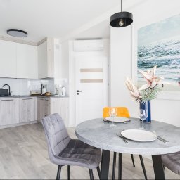 PRZEMYSŁAW JASZCZYK - Jasne, nowoczesne wnętrze: okrągły stół z nakryciem, szare krzesła, kuchnia w tle i plakat z motywem morskim. Aranżacja z kwiatami w wazonie.