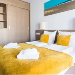 Jasne wnętrze pokoju hotelowego z łóżkiem, żółtą narzutą i dekoracjami ściennymi. Widoczne szafy w zabudowie i lampki nocne. Przytulna aranżacja.