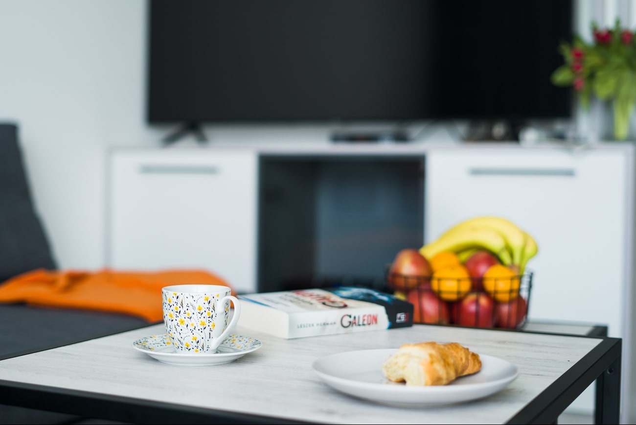 Jasne wnętrze: filiżanka w kwiatki, książka 'Galeon', owoce i rogalik na stoliku kawowym, telewizor w tle. Naturalne światło.