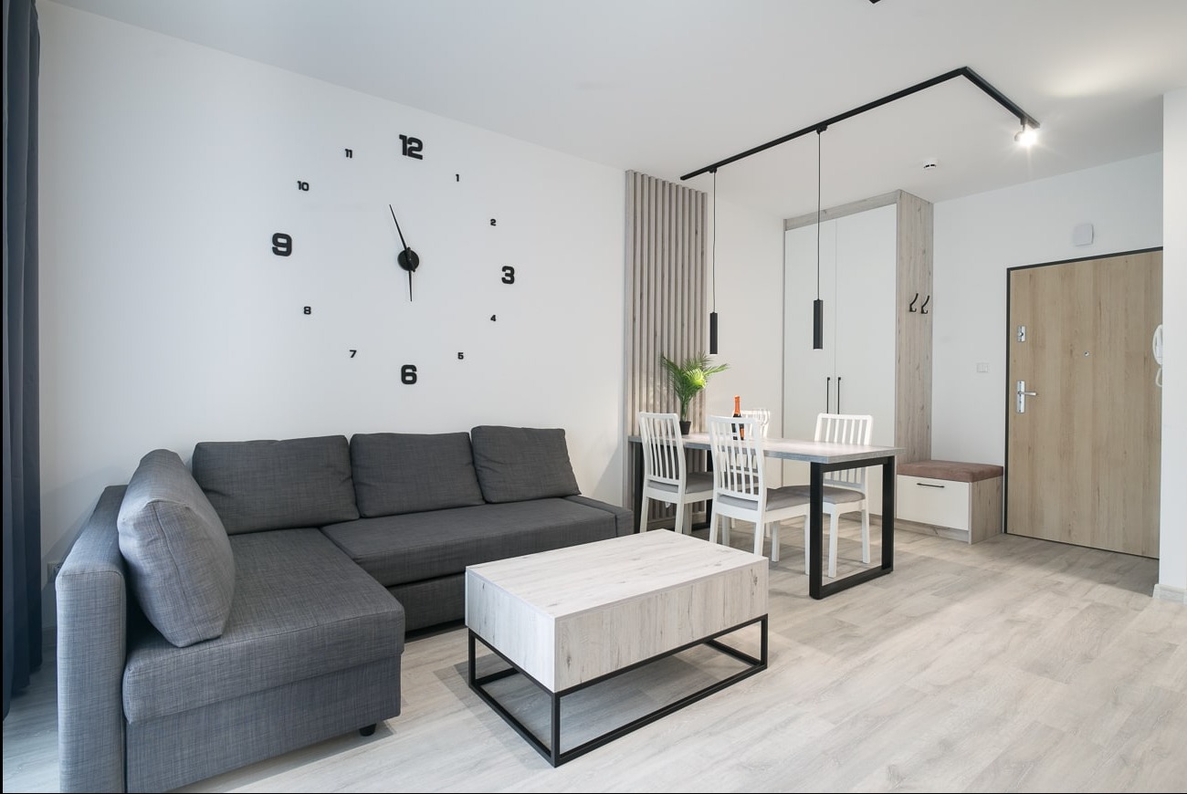 Jasne wnętrze mieszkania z szarą sofą narożną, zegarem ściennym i stołem z krzesłami. Minimalistyczny design, oświetlenie szynowe, drewniana podłoga.