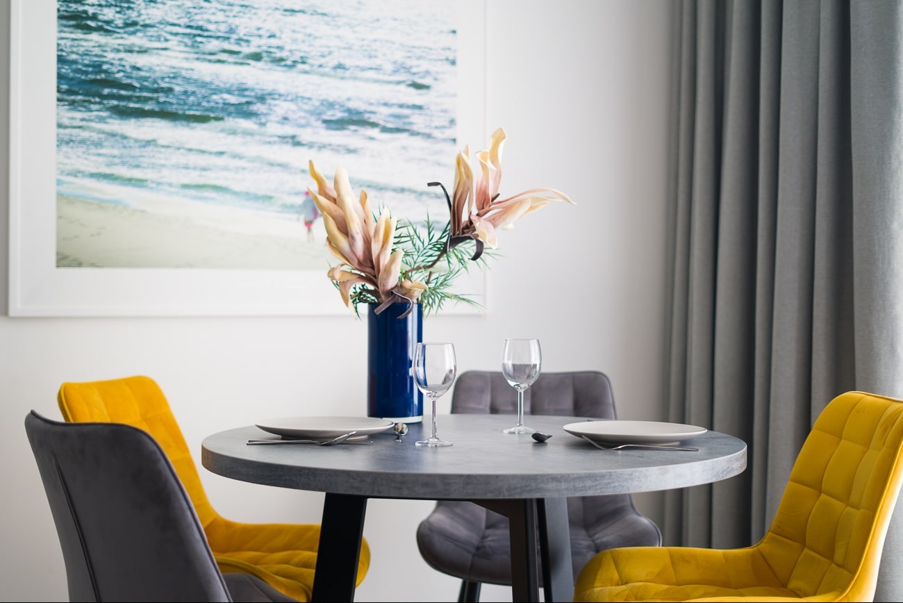 Stylowa jadalnia: okrągły stół z szarego blatu, nakryty dla dwóch osób, otoczony żółtymi i szarymi krzesłami. W tle obraz z motywem morskim i zasłony.