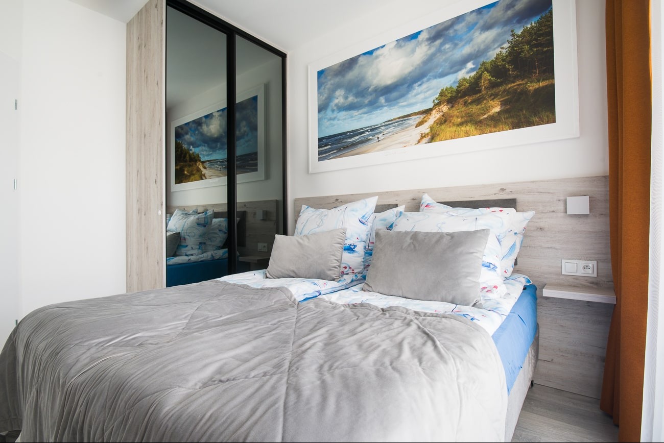 Jasna sypialnia z łóżkiem w szaro-niebieskiej tonacji, nad nim obraz z motywem morza. W tle szafa z lustrzanymi drzwiami.