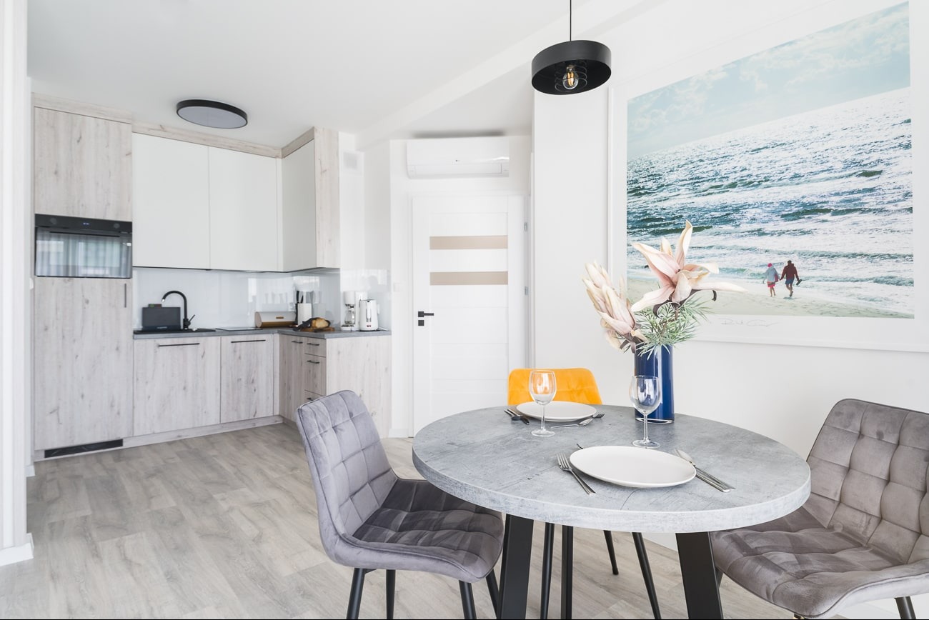 Jasne, nowoczesne wnętrze: okrągły stół z nakryciem, szare krzesła, kuchnia w tle i plakat z motywem morskim. Aranżacja z kwiatami w wazonie.