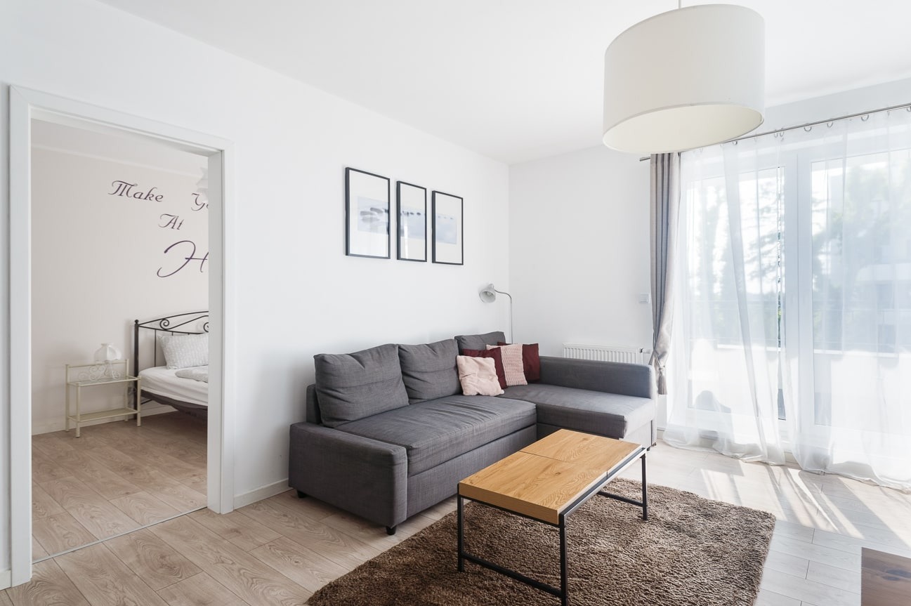 Jasne wnętrze salonu z szarą sofą narożną, drewnianym stolikiem i dekoracjami ściennymi. Widok na sypialnię przez otwarte drzwi. Naturalne światło z okna.