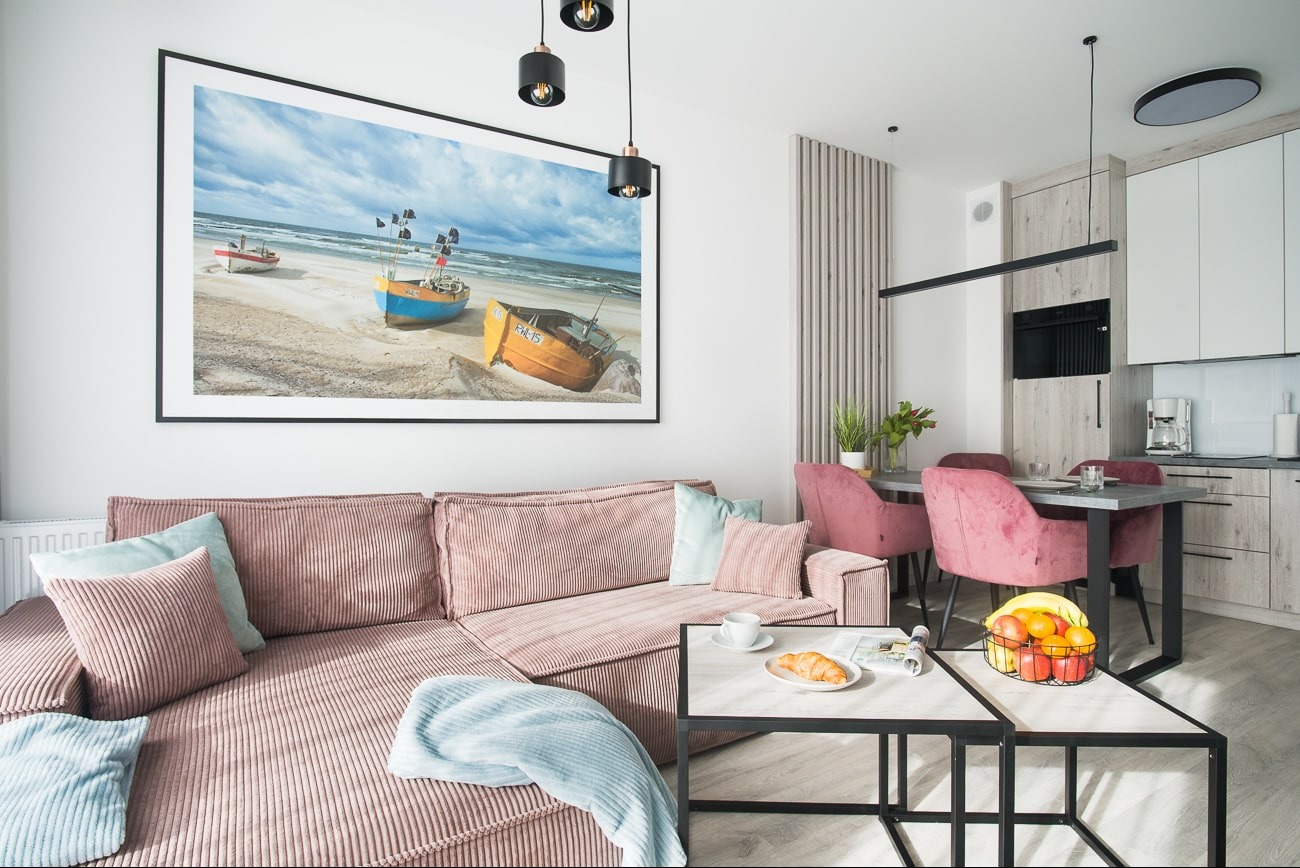 Salon z narożnikiem z różowego sztruksu, stolikami kawowymi i obrazem z motywem rybackich łodzi na ścianie. Widoczna kuchnia i jadalnia w tle.