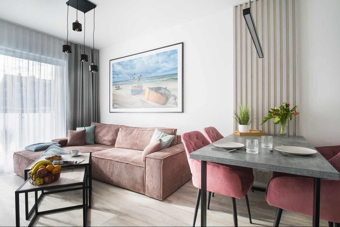 Jasne wnętrze z narożną sofą z różowego sztruksu, stołem z szarym blatem i różowymi krzesłami. Na ścianie wisi obraz z motywem plaży i łodzi.