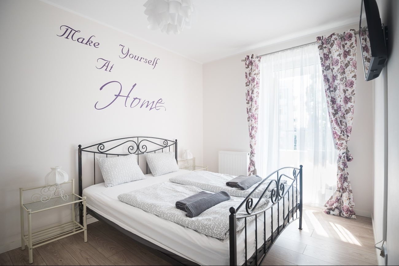 Sypialnia z metalowym łóżkiem, tekstem na ścianie 'Make Yourself At Home', zasłonami w kwiaty i stolikiem nocnym. Jasne, przestronne wnętrze.