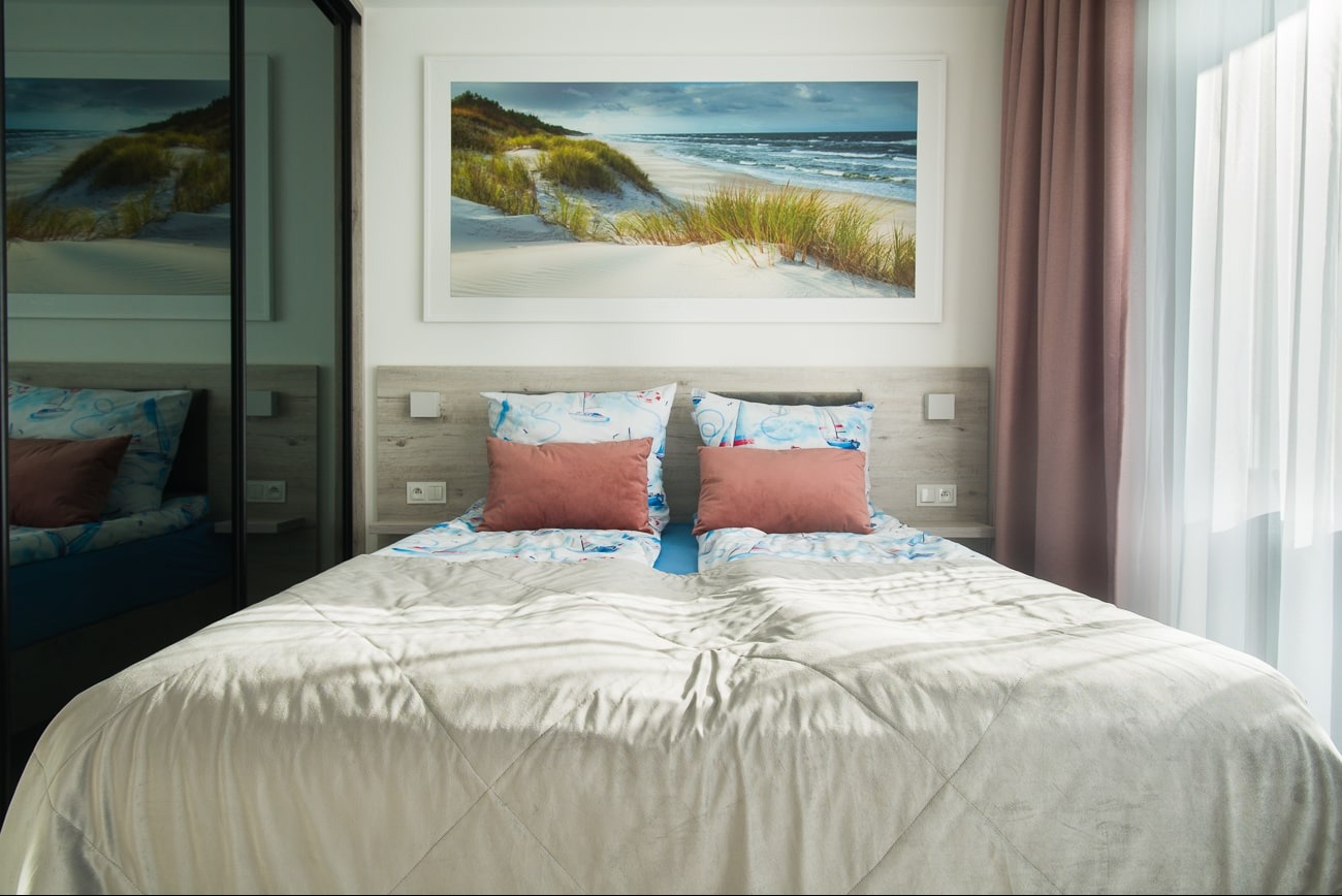 Jasna sypialnia z łóżkiem małżeńskim, nad którym wisi oprawiony obraz z motywem plaży. Po lewej stronie lustrzana szafa, po prawej okno z zasłonami w kolorze brudnego różu.