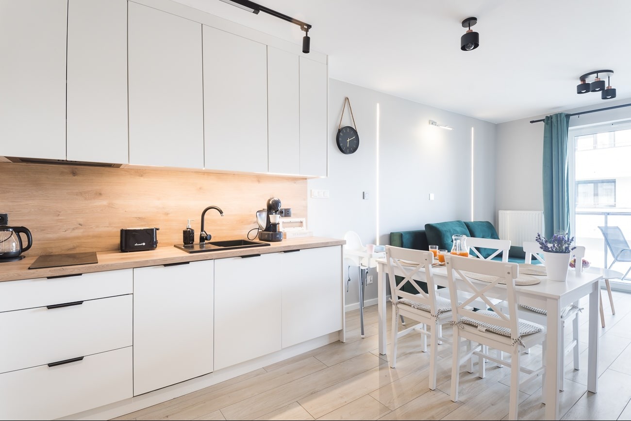 Jasne, nowoczesne wnętrze: biała kuchnia z drewnianym blatem, czarne akcenty, stół z krzesłami i zielona sofa w tle. Minimalistyczny design, ciepłe oświetlenie.
