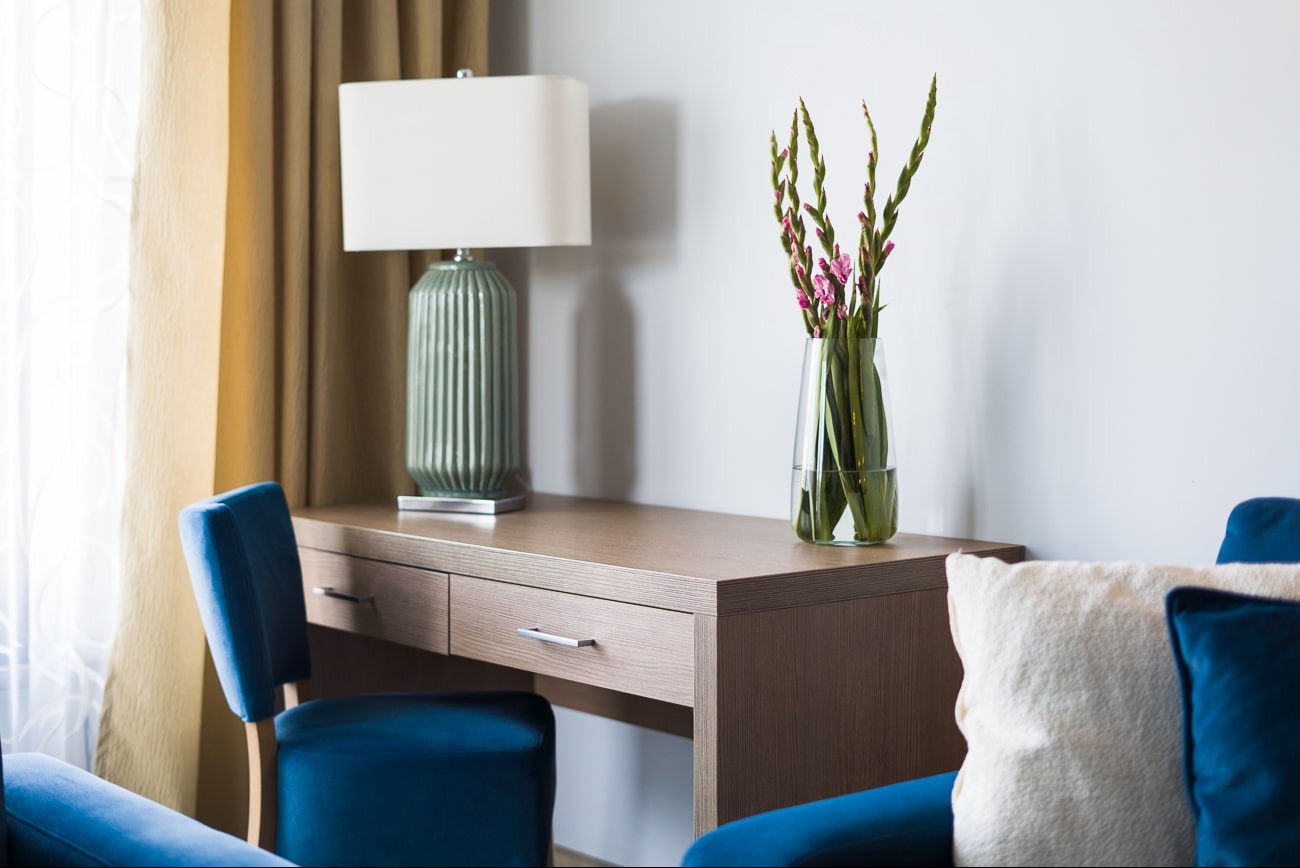 Stylowe biurko z szufladami, lampką nocną i wazonem z kwiatami w jasnym wnętrzu. Niebieskie krzesło i poduszka dodają akcentu kolorystycznego.