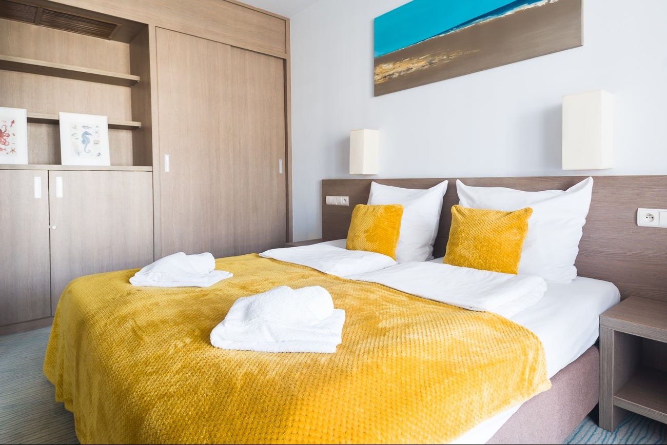 Jasne wnętrze pokoju hotelowego z łóżkiem, żółtą narzutą i dekoracjami ściennymi. Widoczne szafy w zabudowie i lampki nocne. Przytulna aranżacja.