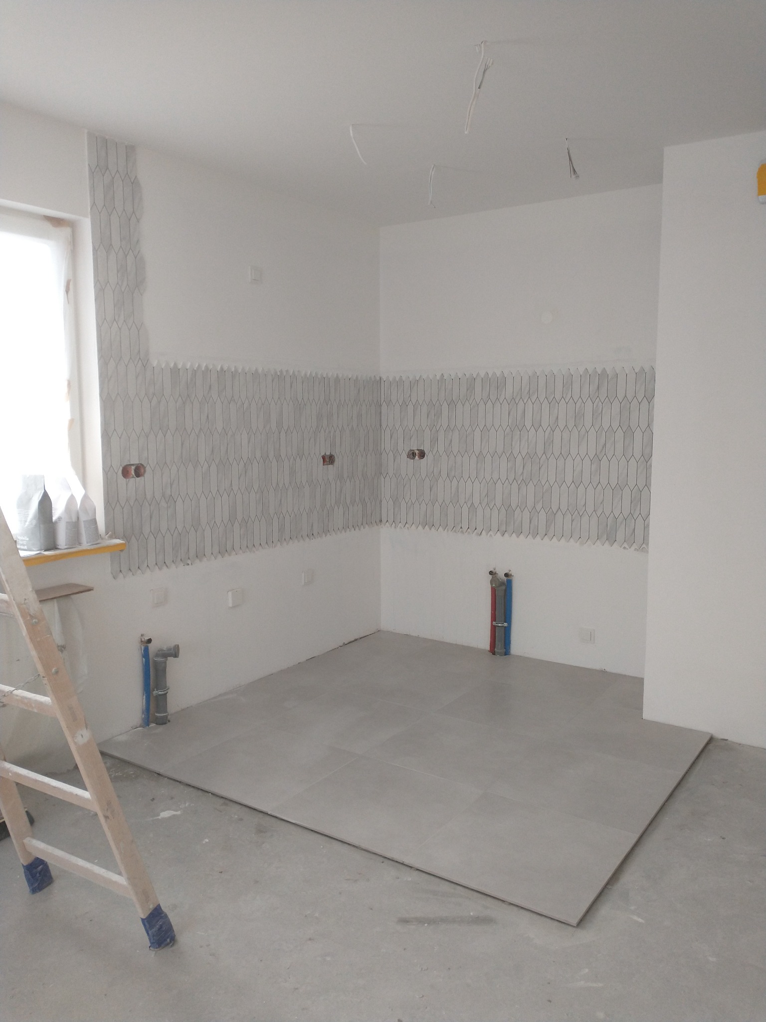 Wnętrze w trakcie prac wykończeniowych: częściowo ułożona mozaika w kształcie rombów na ścianie, szare płytki podłogowe, wystające kable ze ściany i sufitu, drabina oparta o ścianę, worki z zaprawą...