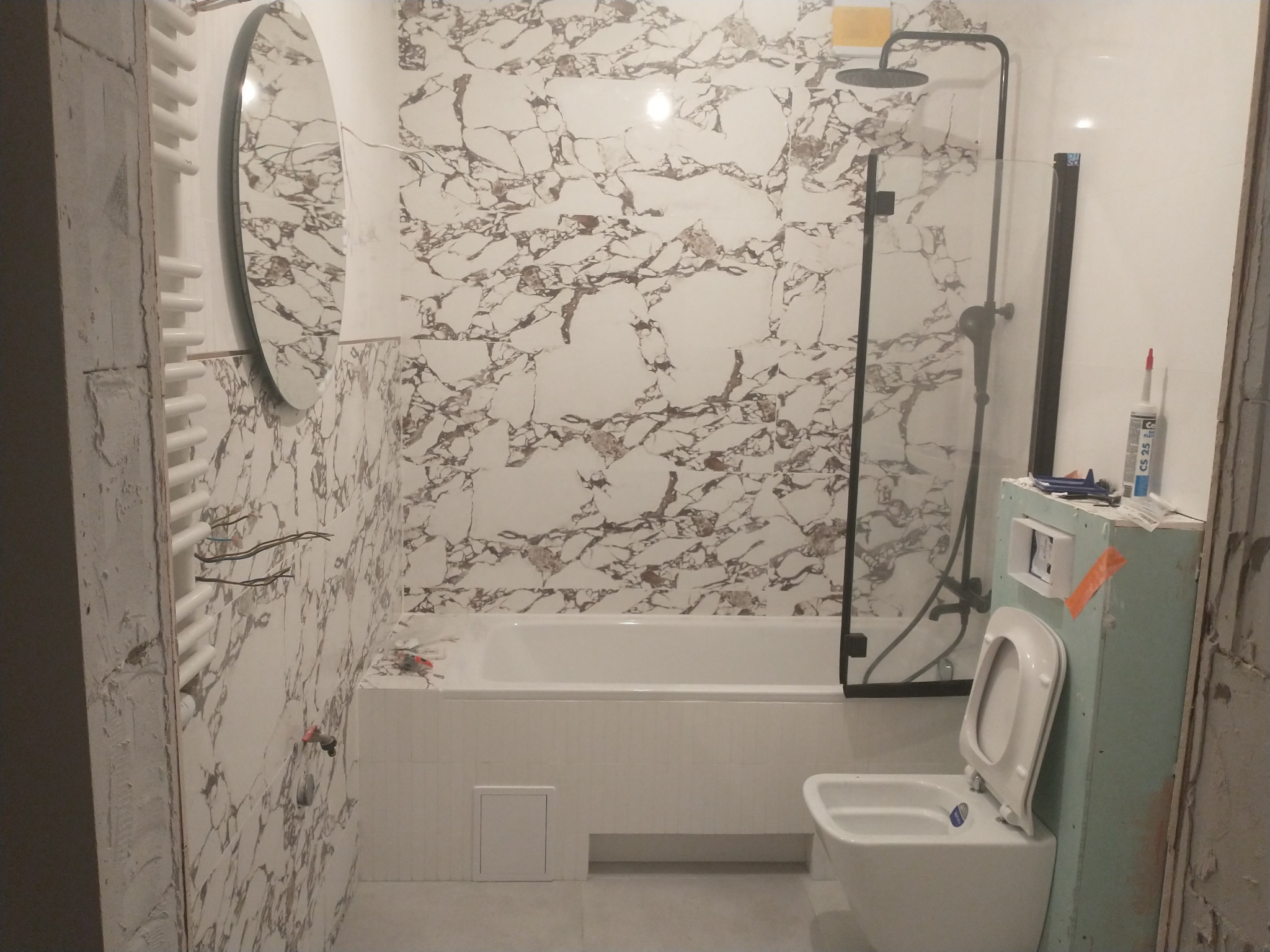 Niedokończona łazienka z wanną obudowaną płytkami, kabiną prysznicową z czarną ramą, wiszącą toaletą i marmurowymi płytkami na ścianach.