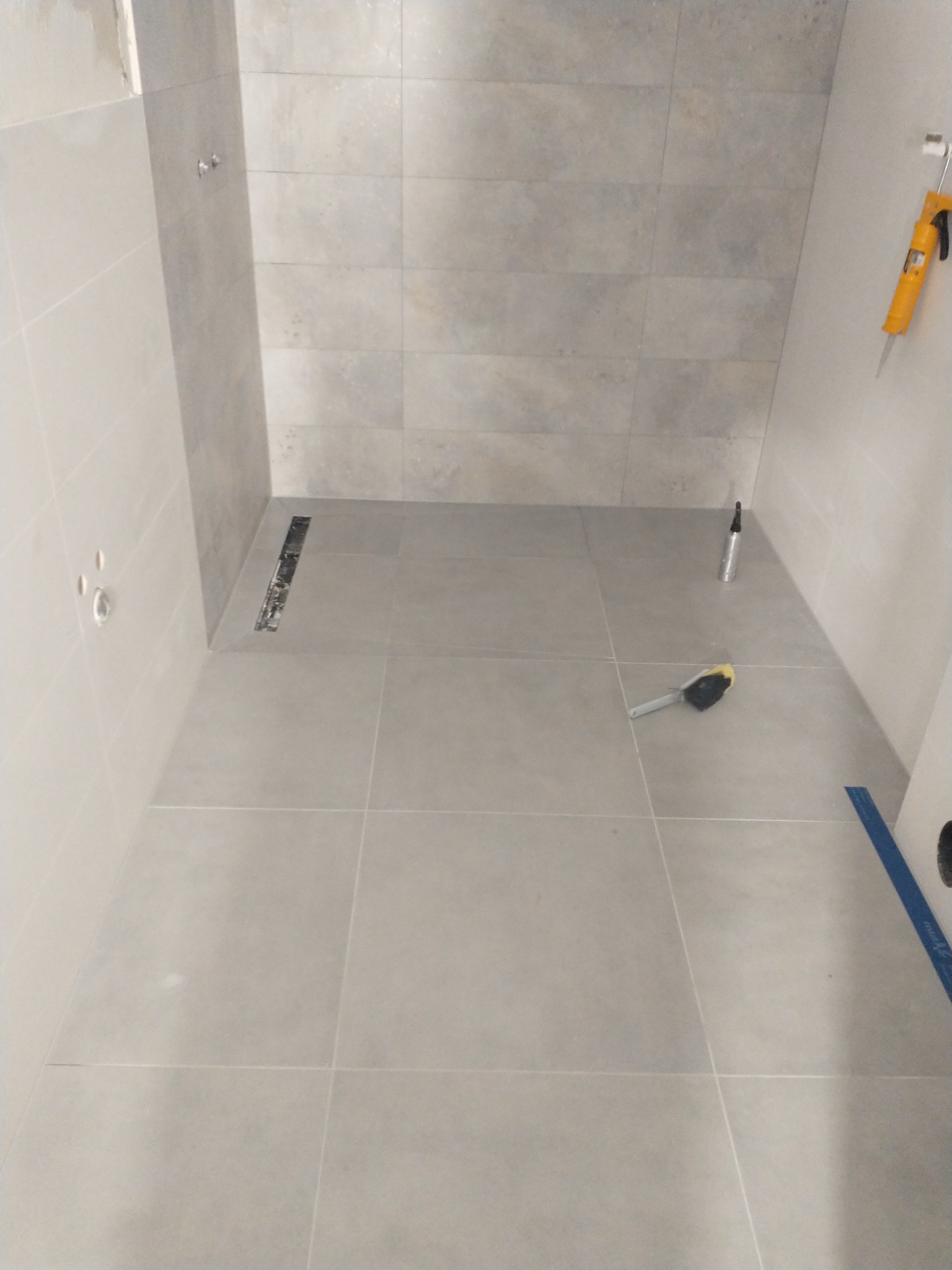 Szara łazienka w trakcie remontu, widoczna listwa odpływowa, pędzel do fugowania, silikon w pistolecie, oraz poziomica, płytki położone na ścianach i podłodze.