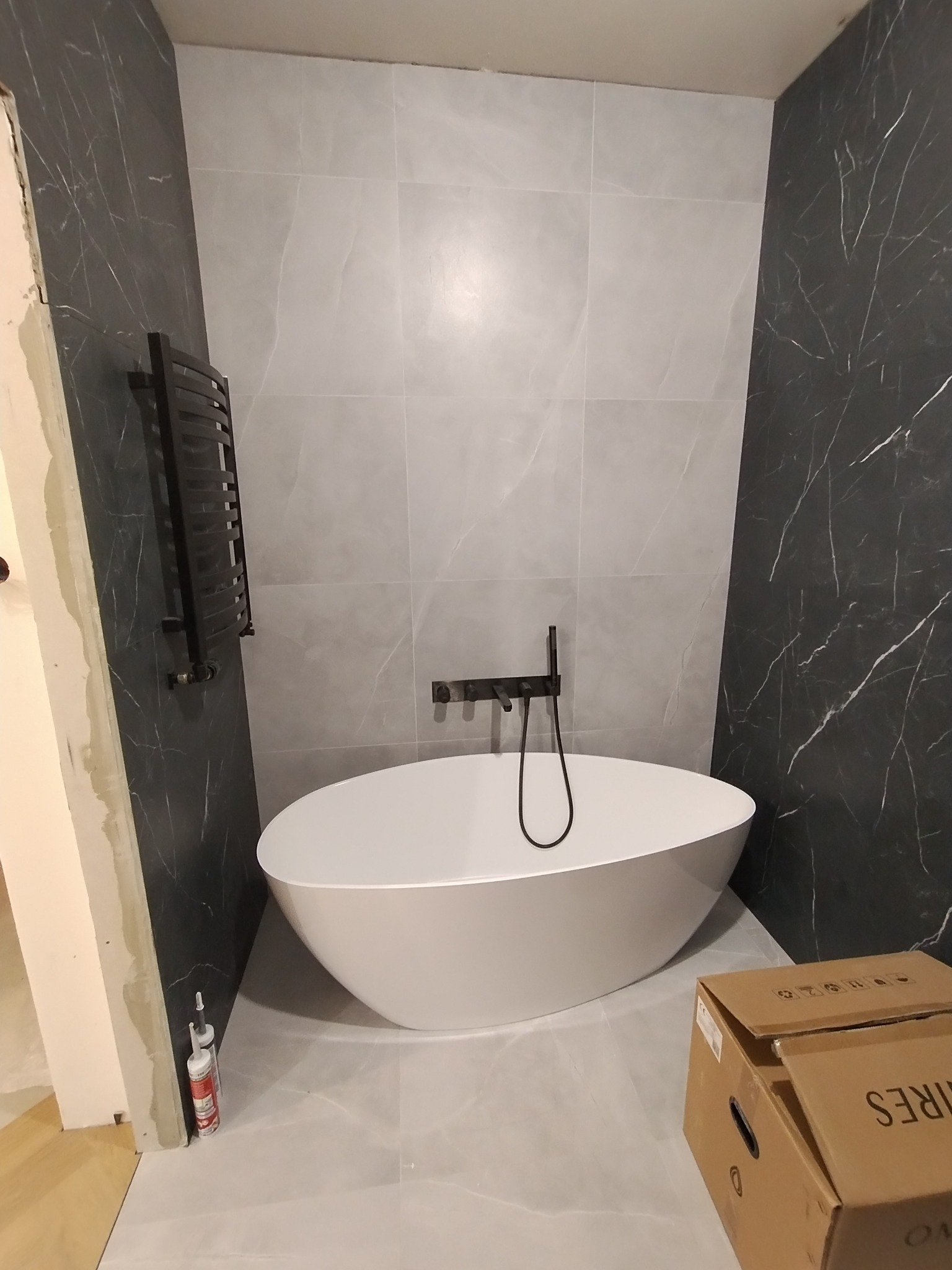 Wanna wolnostojąca o owalnym kształcie z czarną armaturą, otoczona płytkami imitującymi marmur w odcieniach szarości i czerni, z widocznymi tubami silikonu i kartonem na podłodze.