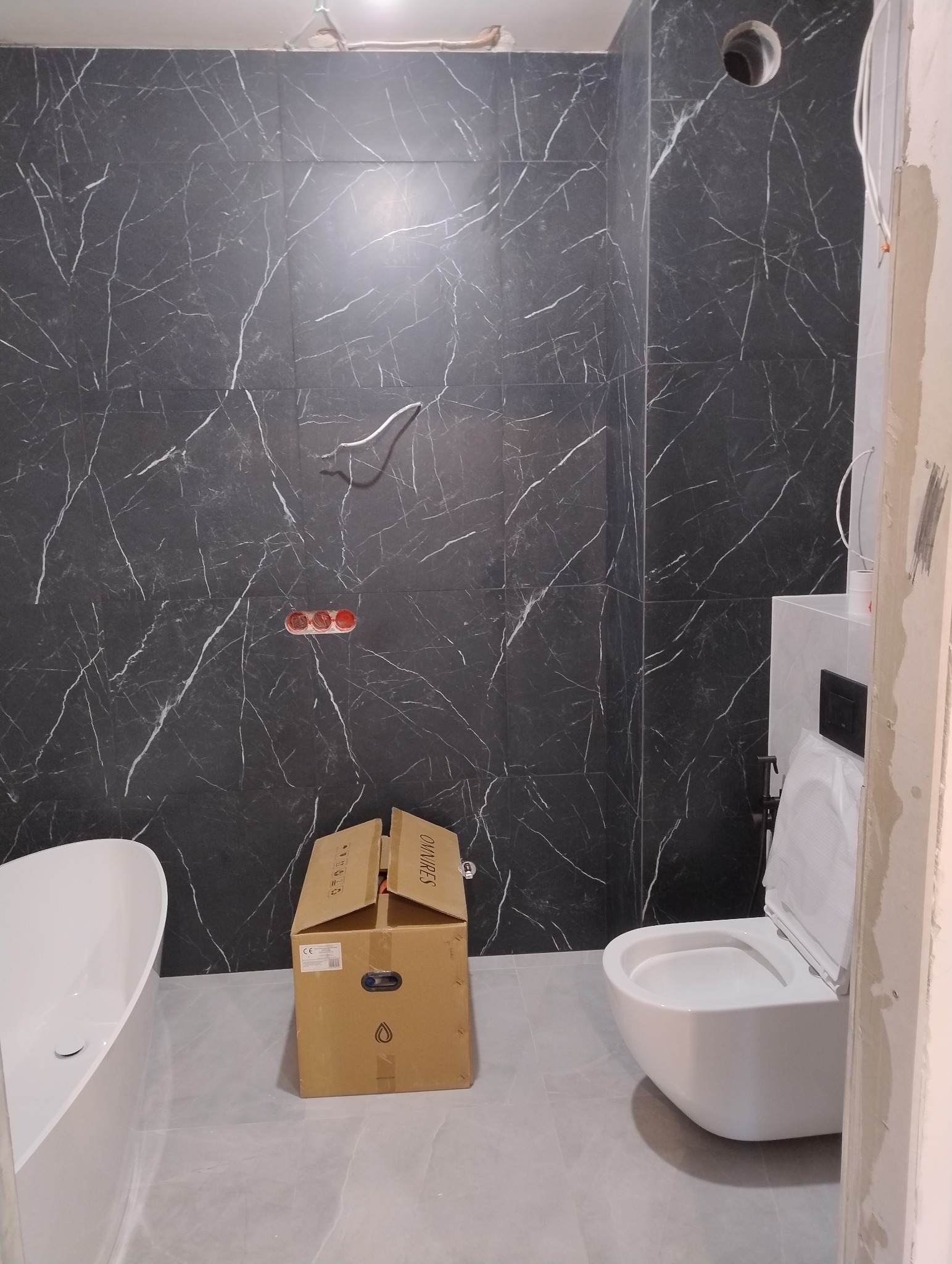 Łazienka w trakcie remontu: czarne płytki imitujące marmur na ścianach, częściowo zamontowana toaleta wisząca, narożnik wanny i karton po armaturze na podłodze.