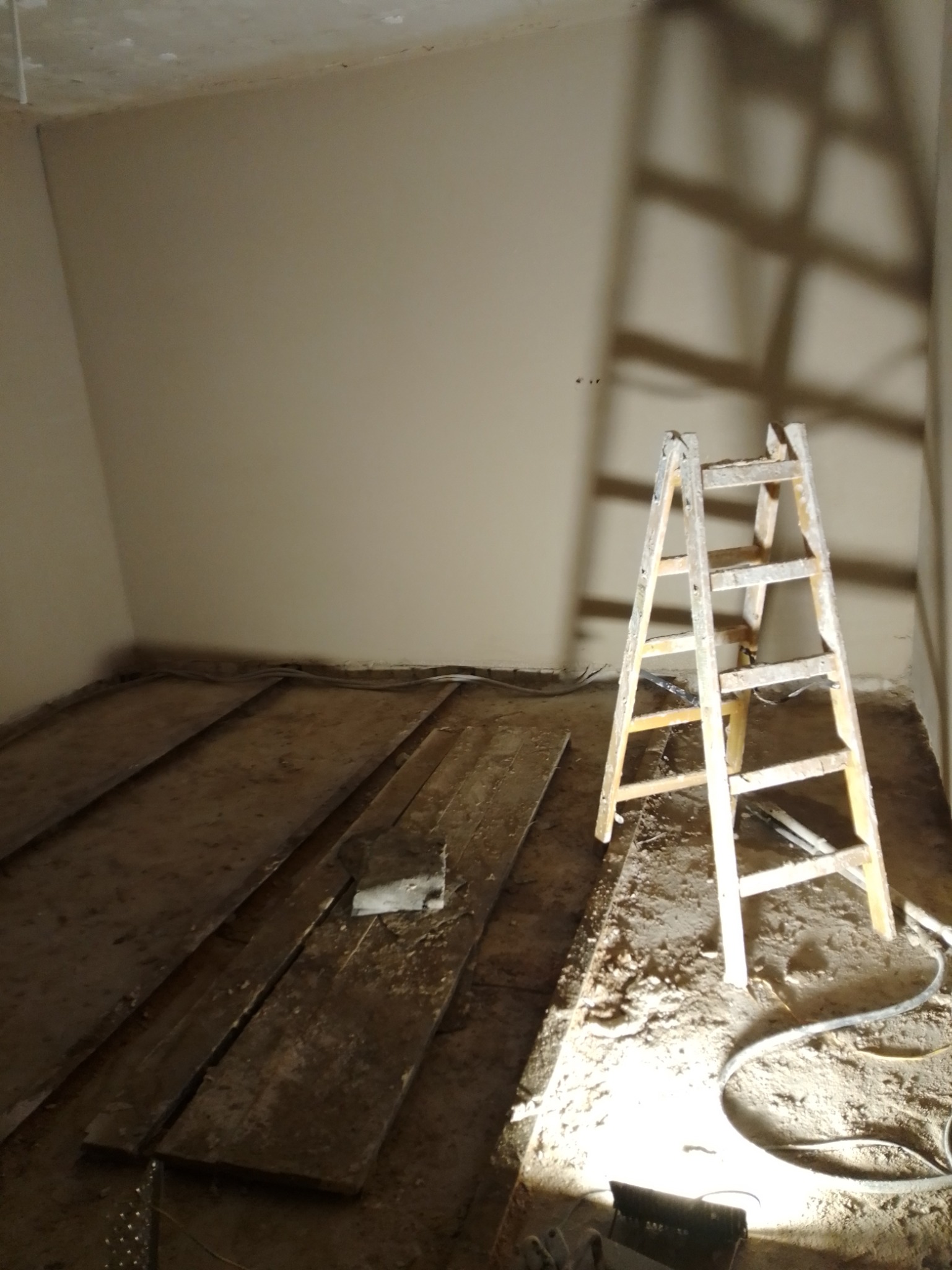Przygotowanie do tynkowania maszynowego: surowe ściany, drewniana podłoga z resztkami gruzu, drabina malarska rzucająca cień na ścianę, widoczne kable i oświetlenie budowlane.