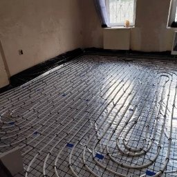 LeoExp Aleksandra Zimowska-Świderska - Białe rury ogrzewania podłogowego ułożone na czarnej siatce w pomieszczeniu z surowymi ścianami, widoczne okno z firanką.