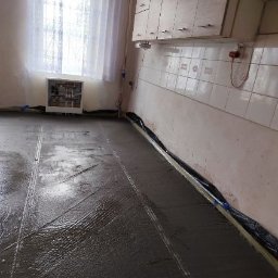 LeoExp Aleksandra Zimowska-Świderska - Wylewka betonowa na podłodze w kuchni, widoczne listwy dylatacyjne przy ścianach, okno z firanką w tle.