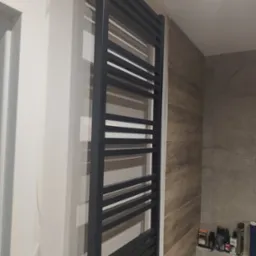 Zamontowany, czarny grzejnik łazienkowy drabinkowy na ścianie wyłożonej płytkami imitującymi drewno. Widoczne kosmetyki na półce. Świeżo wykończona łazienka.