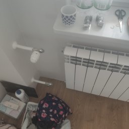 LeoExp Aleksandra Zimowska-Świderska - Biały grzejnik panelowy zamontowany przy ścianie, widoczne rury doprowadzające wodę, termostat, oraz elementy instalacyjne na parapecie okiennym.