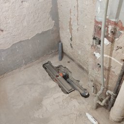 LeoExp Aleksandra Zimowska-Świderska - Szczegółowe ujęcie odpływu liniowego w surowej łazience przed wykończeniem. Widoczne rury, surowe ściany i podłoga w trakcie prac hydraulicznych.