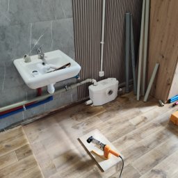 LeoExp Aleksandra Zimowska-Świderska - Biała umywalka z baterią na tle szarej płytki, obok biały zbiornik i rury. Na podłodze narzędzie do spawania rur na desce. Widoczne etapy prac hydraulicznych.