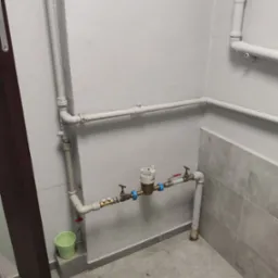 Zamontowana instalacja wodna z licznikiem w pomieszczeniu z szarą glazurą i rurami pomalowanymi na biało. Widoczny zawór i zielone wiaderko.