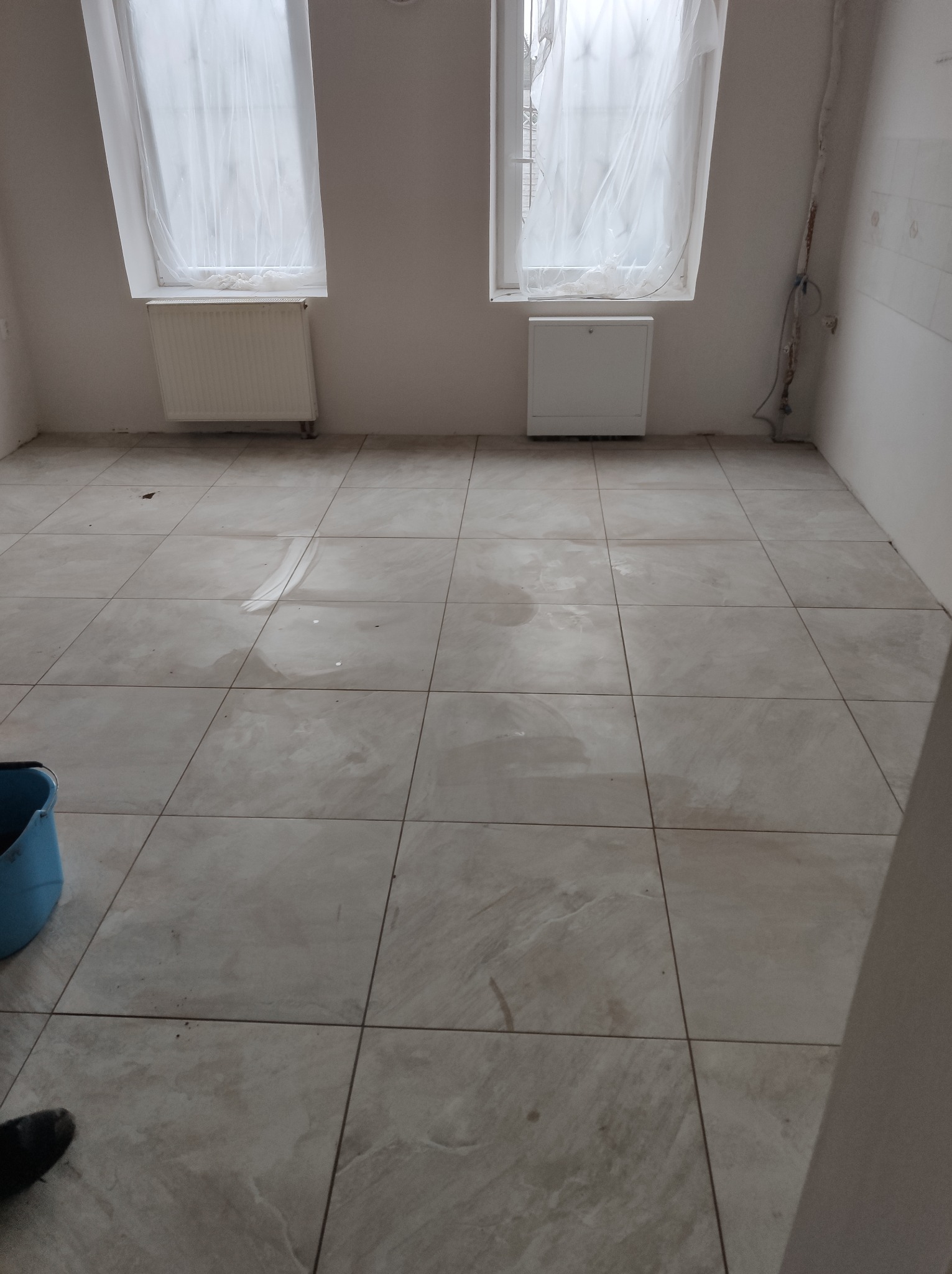 Wykończone płytki podłogowe w jasnym odcieniu w pomieszczeniu z grzejnikami pod oknami, widoczny fragment niebieskiego wiadra i buta.