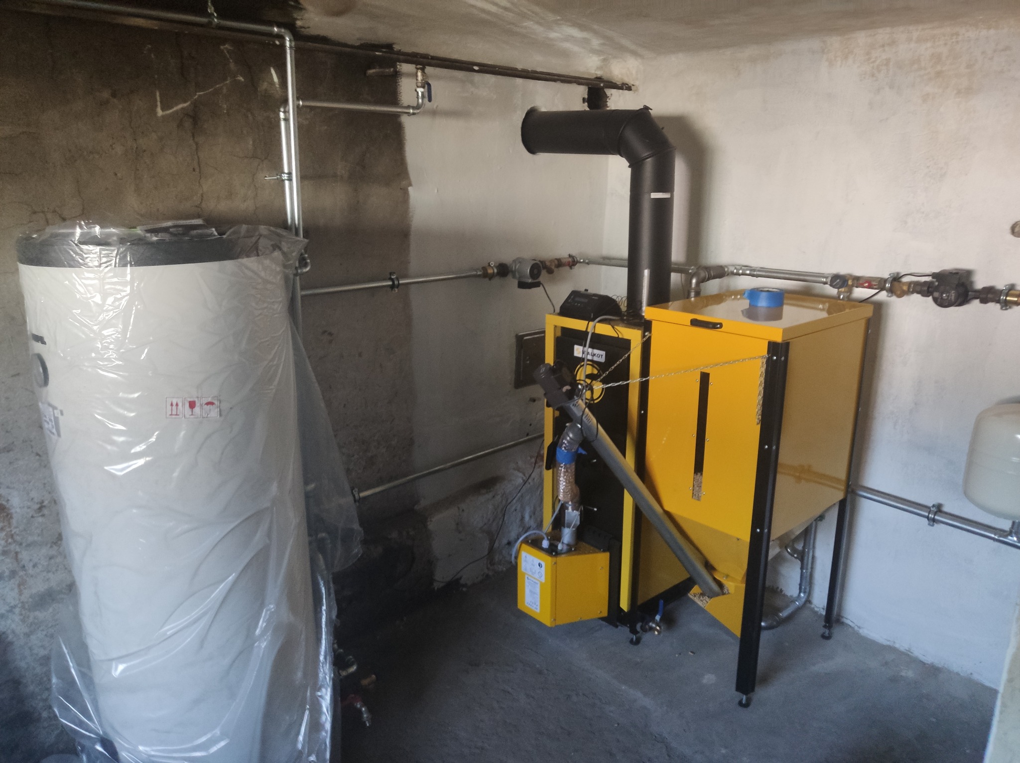 Żółty piec CO na paliwo stałe z czarnym kominem, obok biały zbiornik na wodę w folii ochronnej, widoczne rury instalacji grzewczej na tle surowych ścian piwnicy.