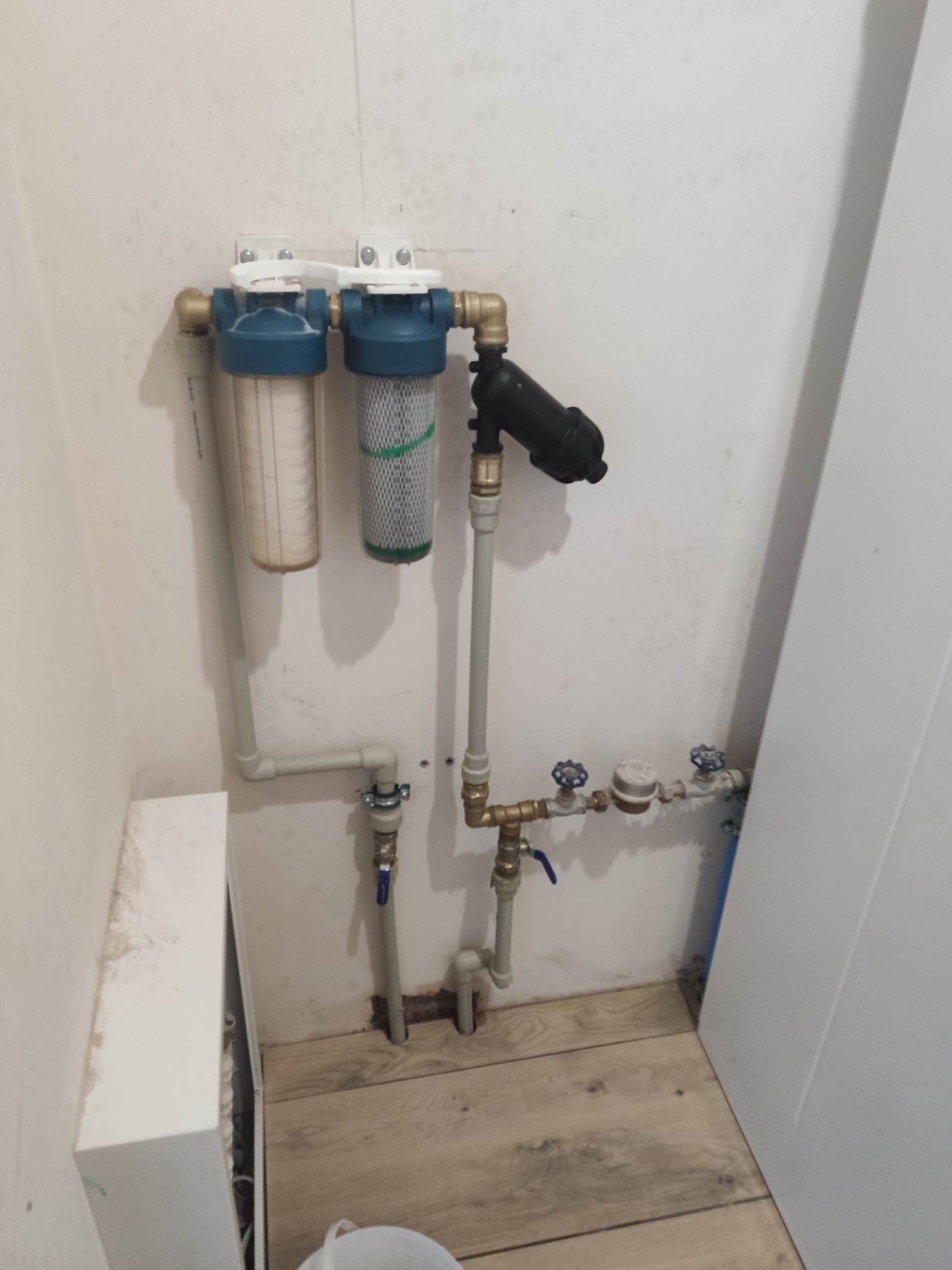 Zamontowany system filtracji wody z rurami i zaworami, widoczny na tle białej ściany i drewnopodobnej podłogi. Filtr dwuczęściowy z elementami w kolorze niebieskim.