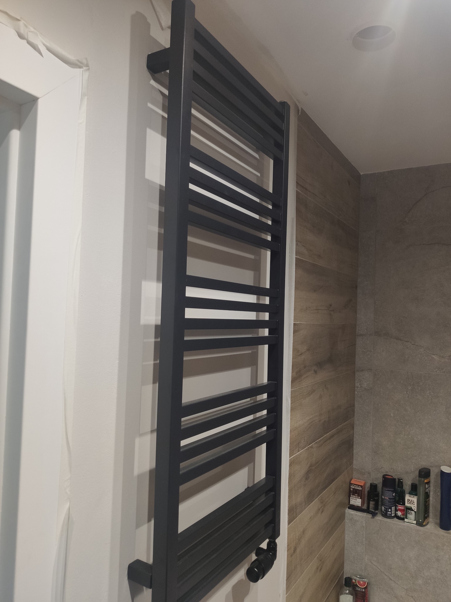 Zamontowany, czarny grzejnik łazienkowy drabinkowy na ścianie wyłożonej płytkami imitującymi drewno. Widoczne kosmetyki na półce. Świeżo wykończona łazienka.