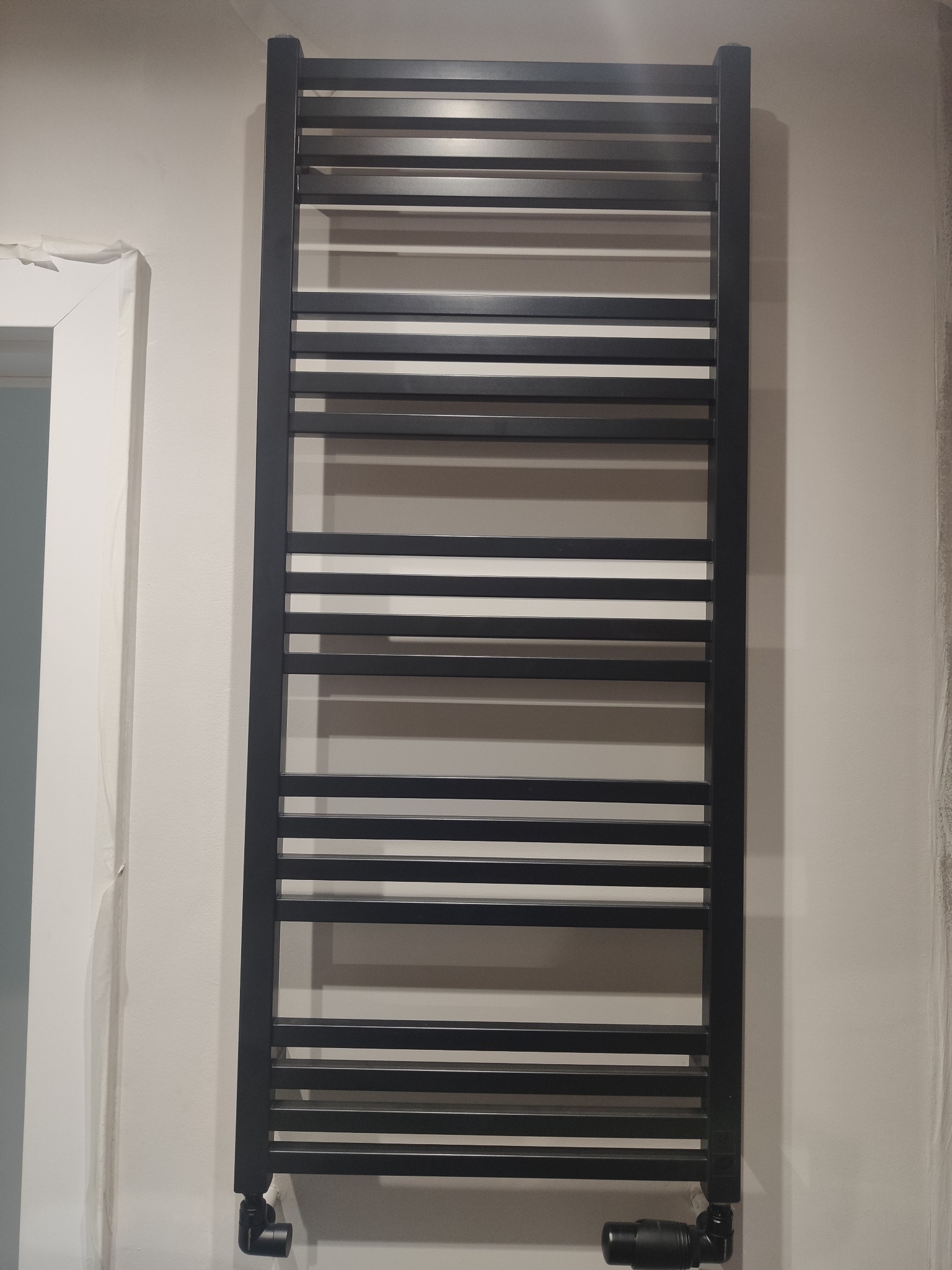 Czarny, drabinkowy grzejnik łazienkowy na ścianie, z widocznymi zaworami, na tle jasnej ściany i fragmentu okna.