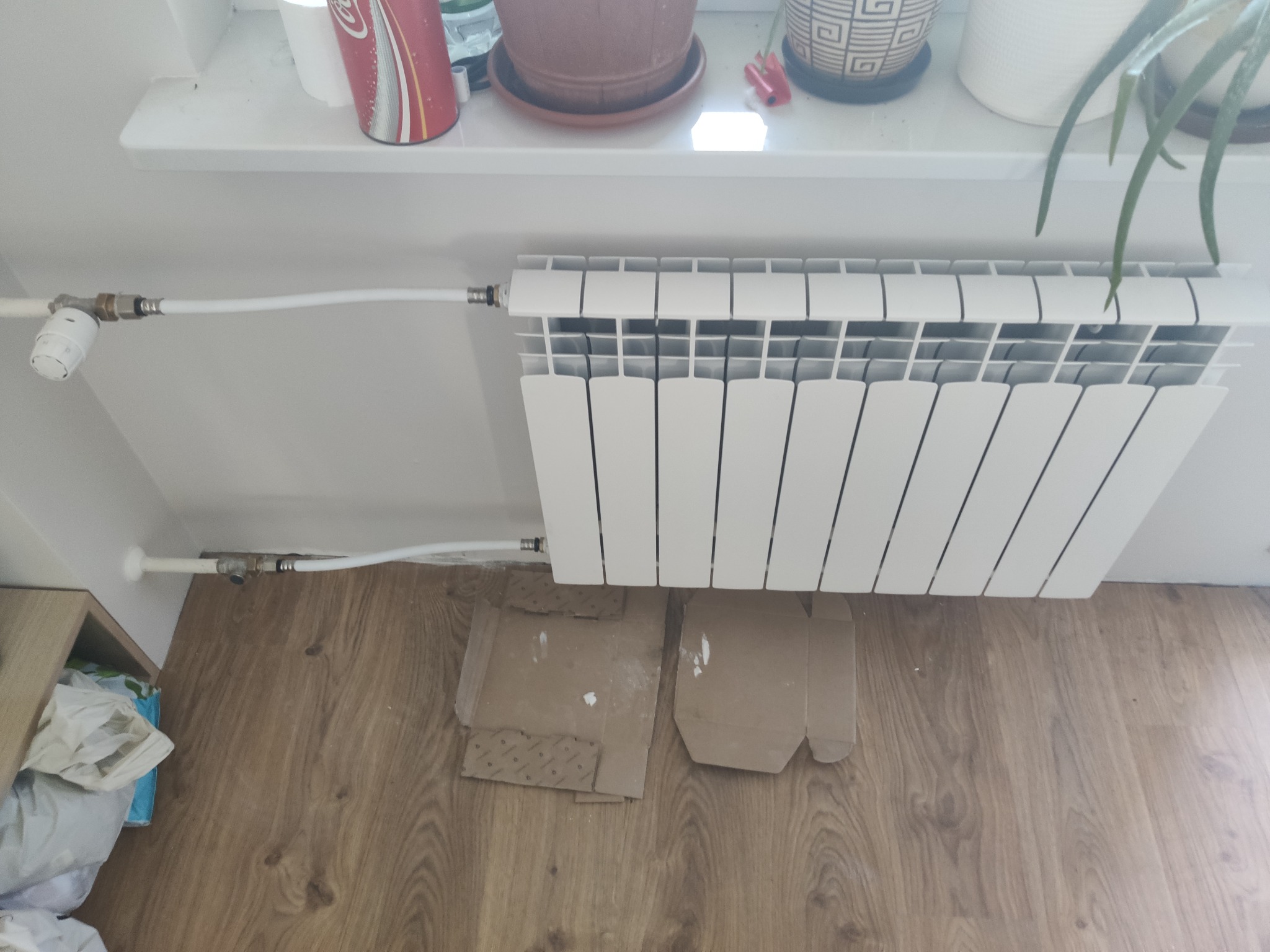 Biały, nowy grzejnik panelowy z termostatem zamontowany na ścianie, z widocznymi białymi rurami doprowadzającymi, na podłodze kartony zabezpieczające.