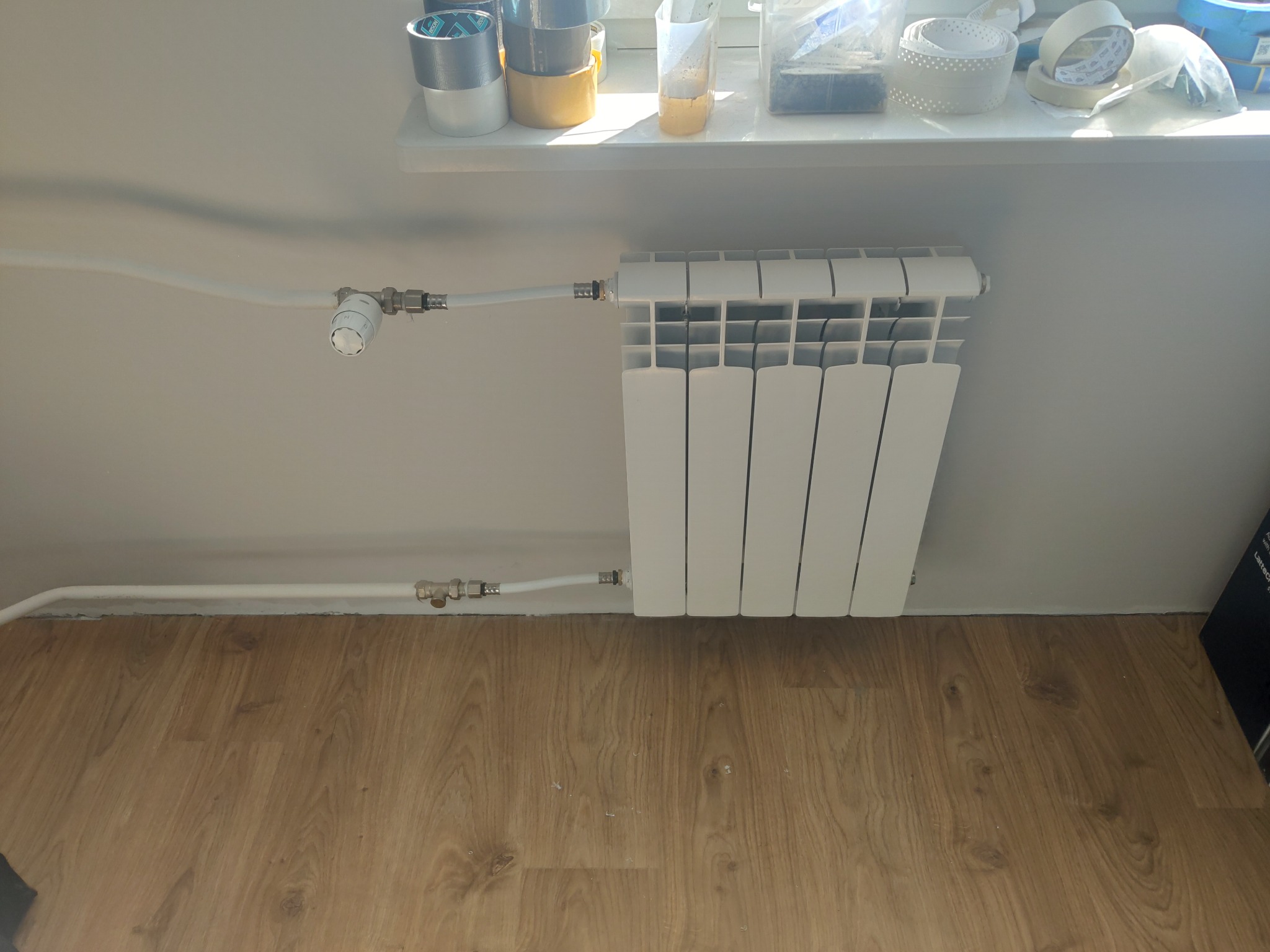 Biały grzejnik panelowy zamontowany przy ścianie, z widocznymi rurami doprowadzającymi i termostatem, na tle drewnianej podłogi i parapetu z akcesoriami.