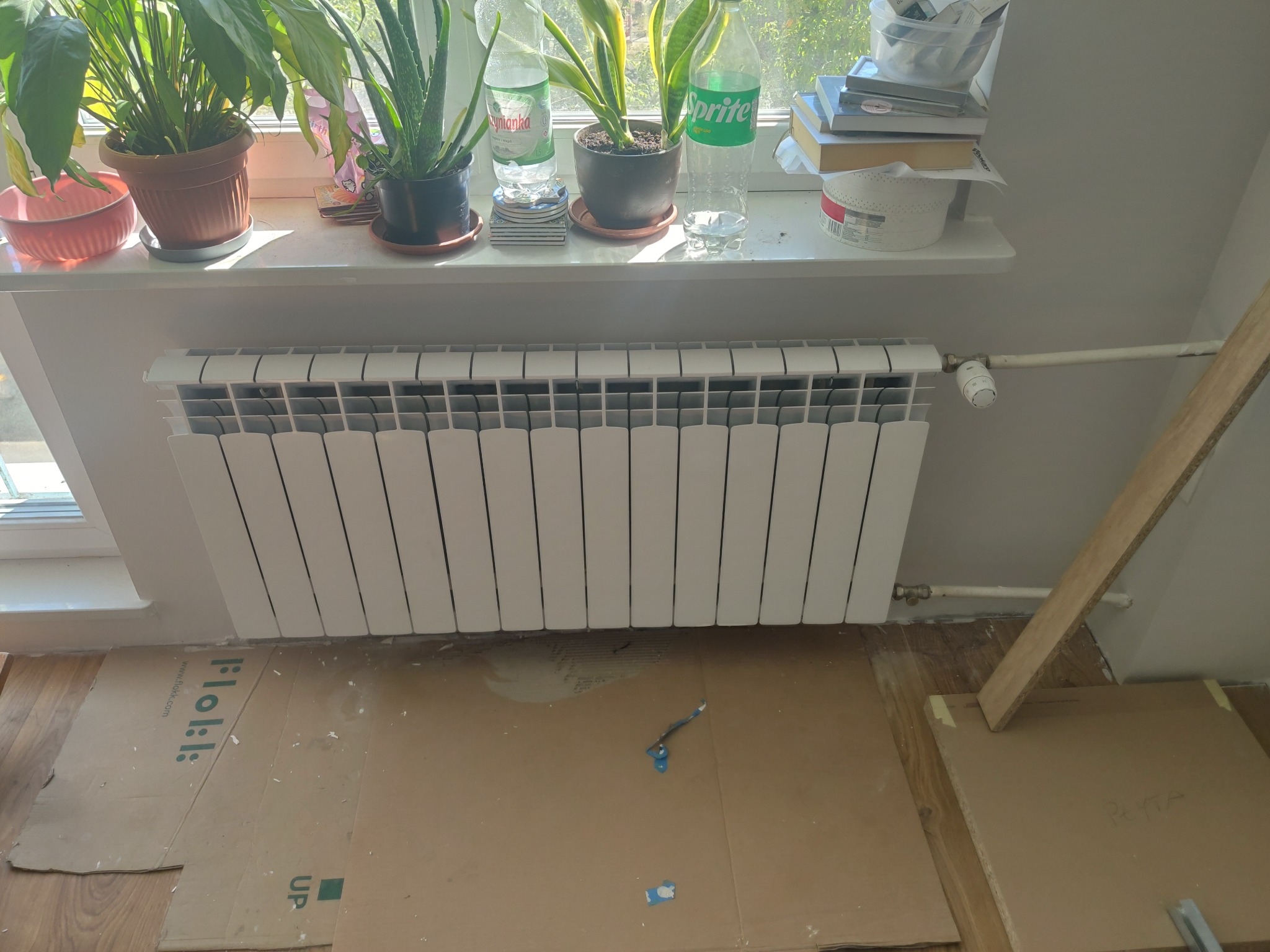 Biały, nowy grzejnik panelowy zamontowany pod oknem na tle kartonów zabezpieczających podłogę. Na parapecie doniczki z roślinami i butelki z napojami.