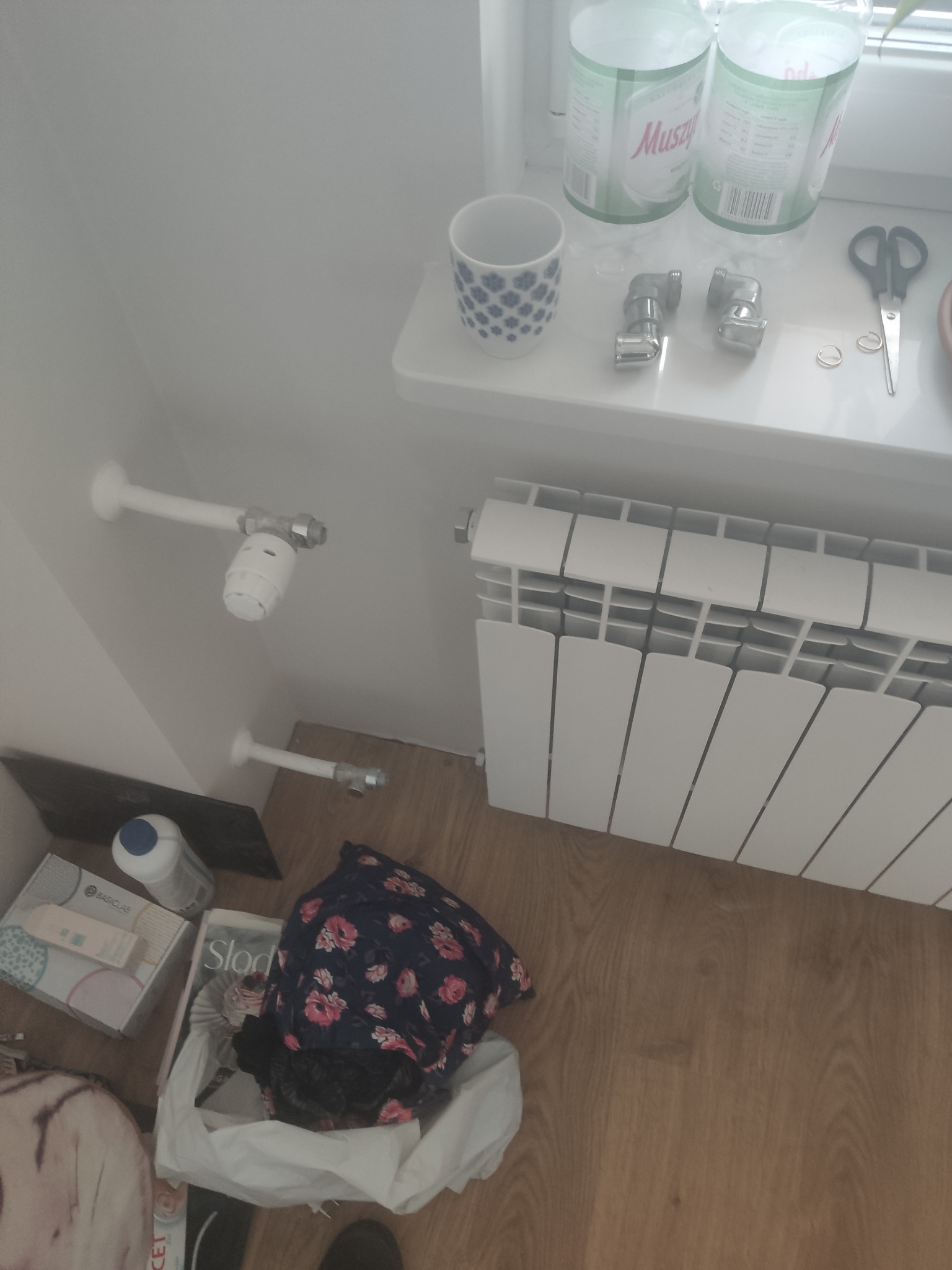 Biały grzejnik panelowy zamontowany przy ścianie, widoczne rury doprowadzające wodę, termostat, oraz elementy instalacyjne na parapecie okiennym.