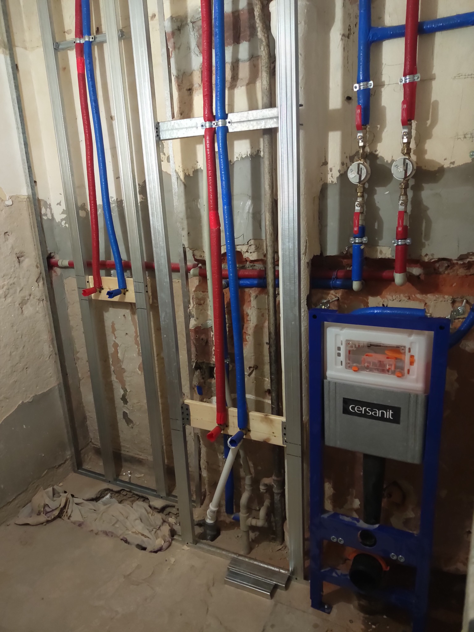 Instalacja wodna z rurami w kolorach czerwonym i niebieskim, zamontowana na metalowym stelażu przy ścianie z odsłoniętą cegłą i elementami instalacji podtynkowych Cersanit.