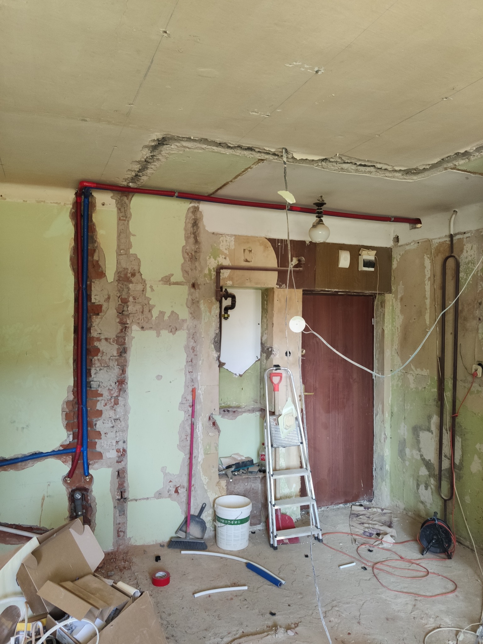 Wnętrze w trakcie remontu: odsłonięte cegły, nowe rury instalacji w kolorze czerwonym i niebieskim. Widoczna drabina, narzędzia i pozostałości po skuciu tynku.