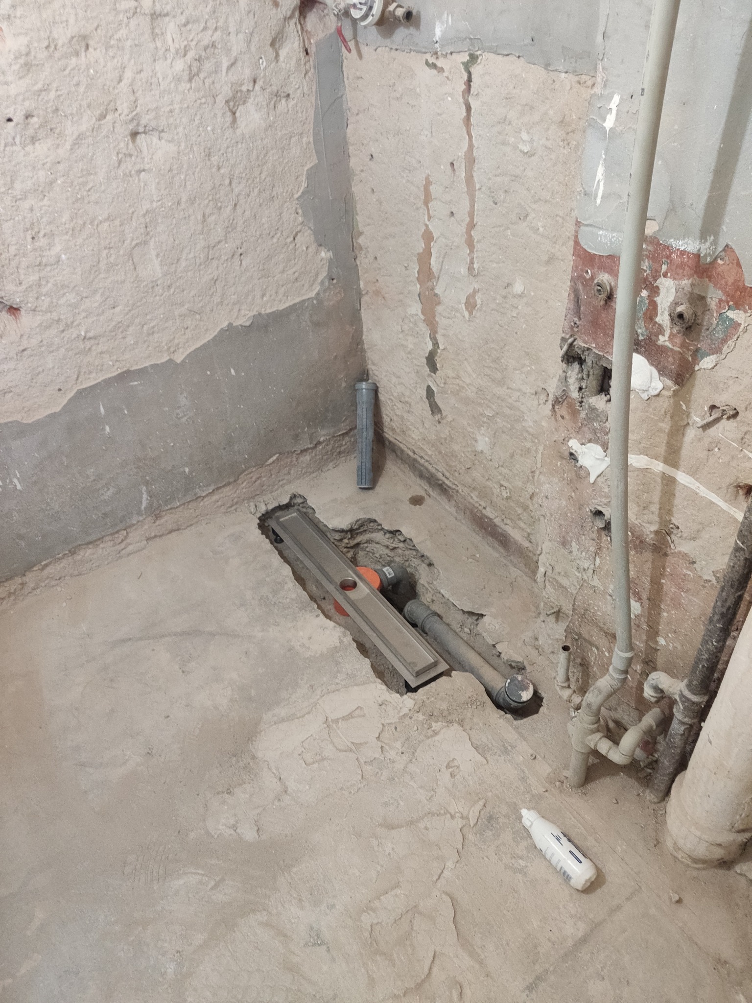 Szczegółowe ujęcie odpływu liniowego w surowej łazience przed wykończeniem. Widoczne rury, surowe ściany i podłoga w trakcie prac hydraulicznych.