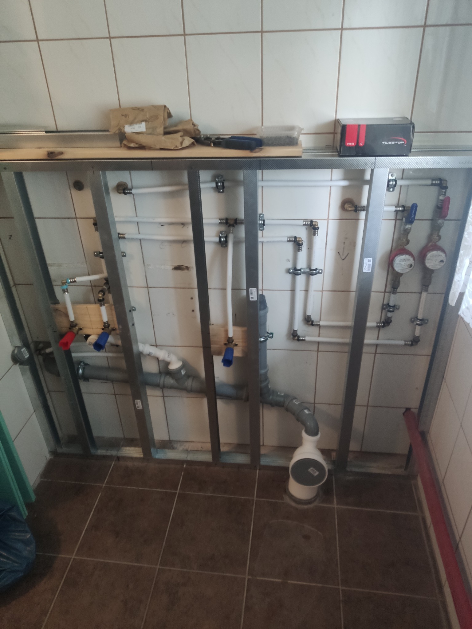 Instalacja wodno-kanalizacyjna w trakcie montażu, rury z tworzywa i metalu, liczniki wody, stelaż podtynkowy, płytki ceramiczne w tle.