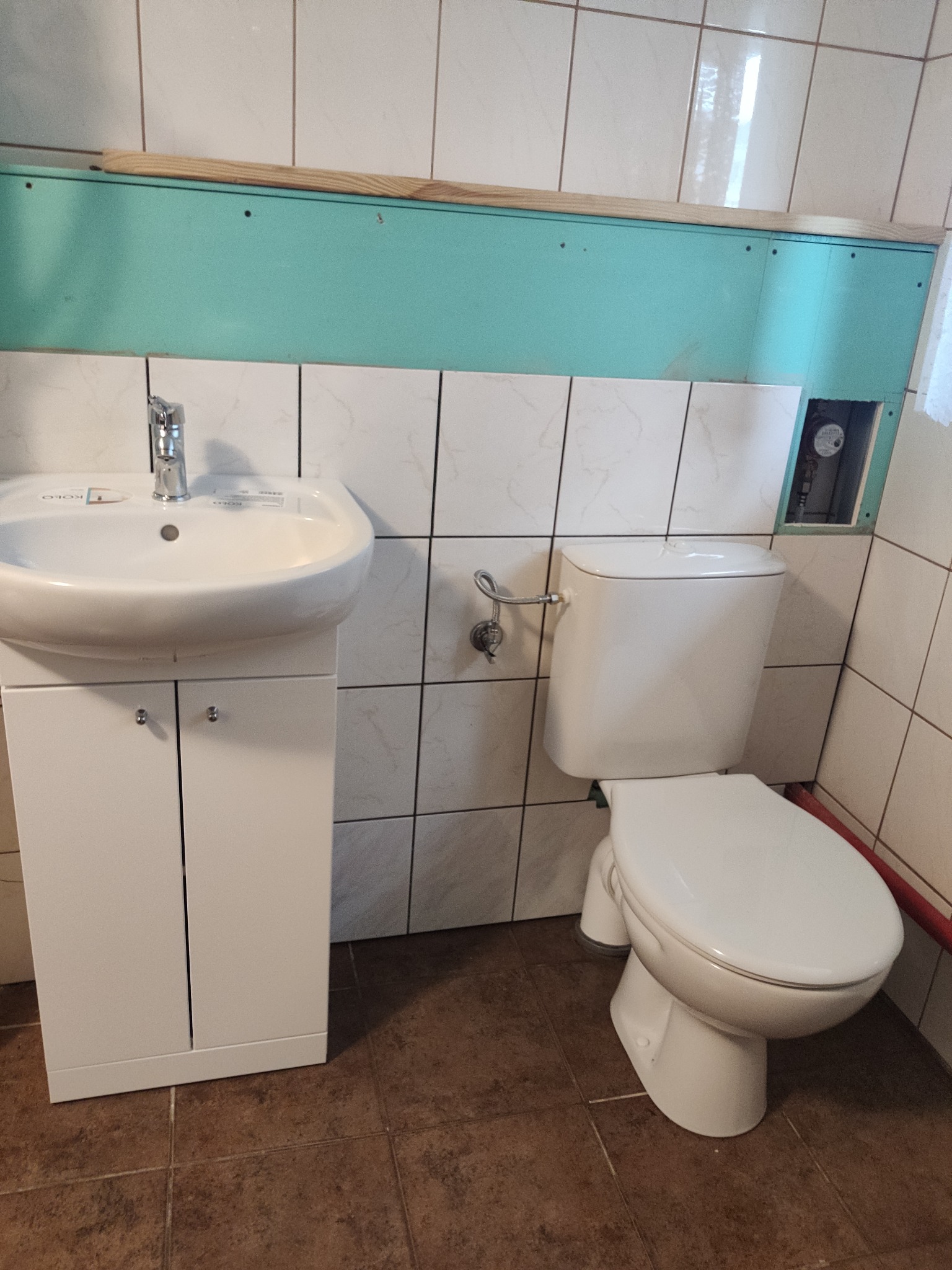 Biała umywalka z szafką i toaleta w łazience z białymi i brązowymi płytkami. Widoczna instalacja za zabudową.