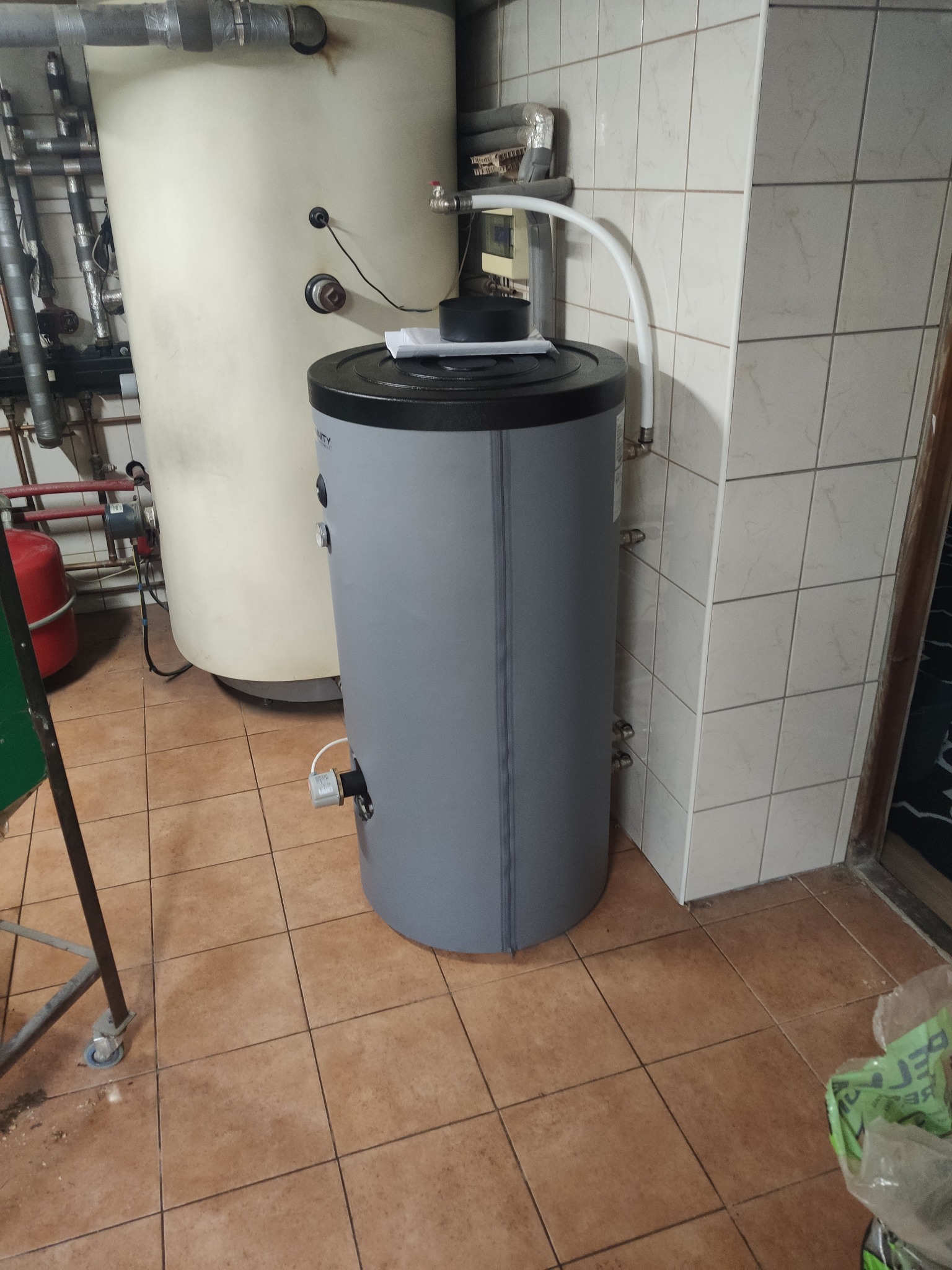 Szary, cylindryczny bojler z czarną górą stoi w rogu pomieszczenia z beżowymi płytkami. W tle widoczny kremowy zbiornik i elementy instalacji.