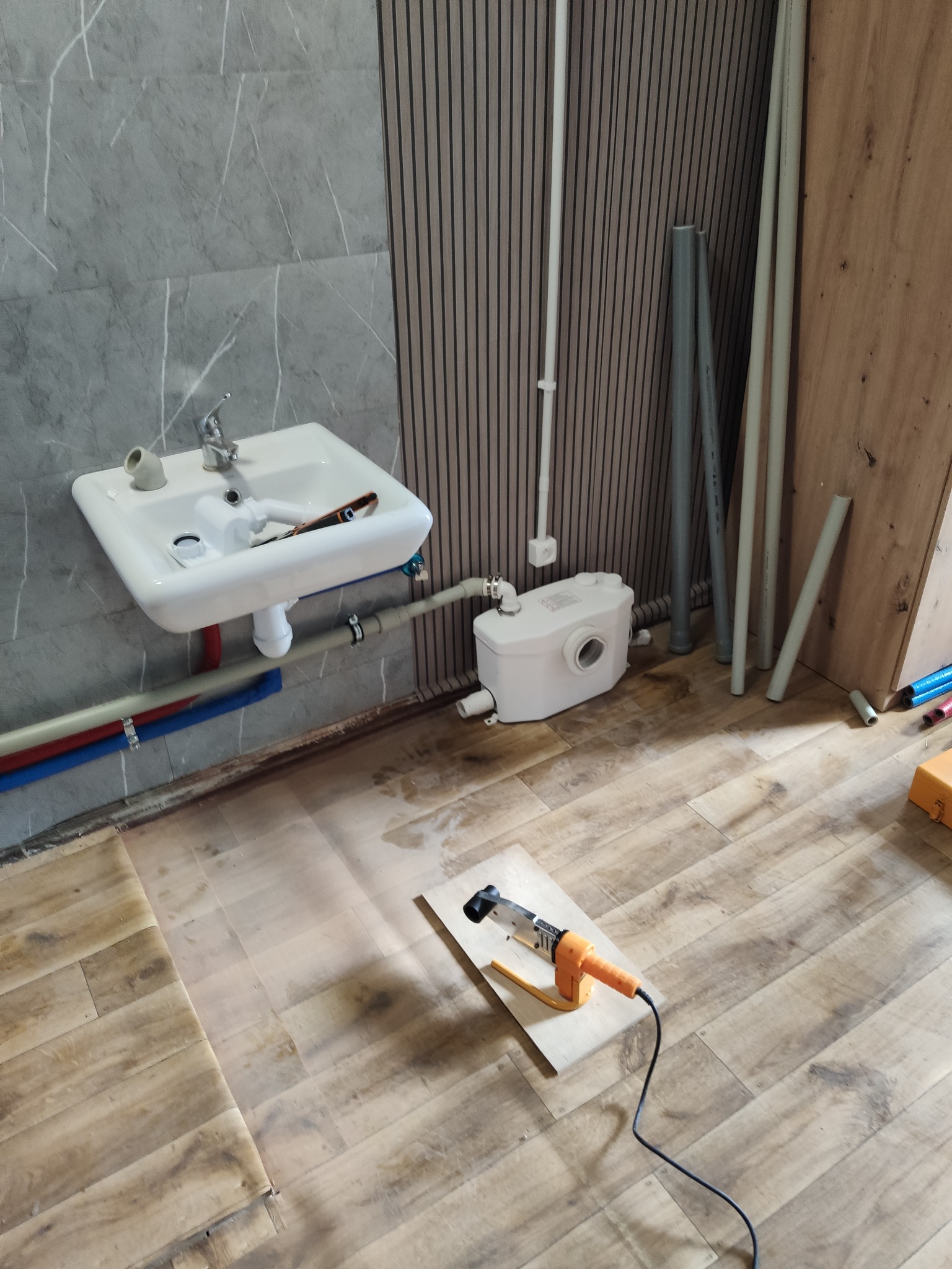 Biała umywalka z baterią na tle szarej płytki, obok biały zbiornik i rury. Na podłodze narzędzie do spawania rur na desce. Widoczne etapy prac hydraulicznych.