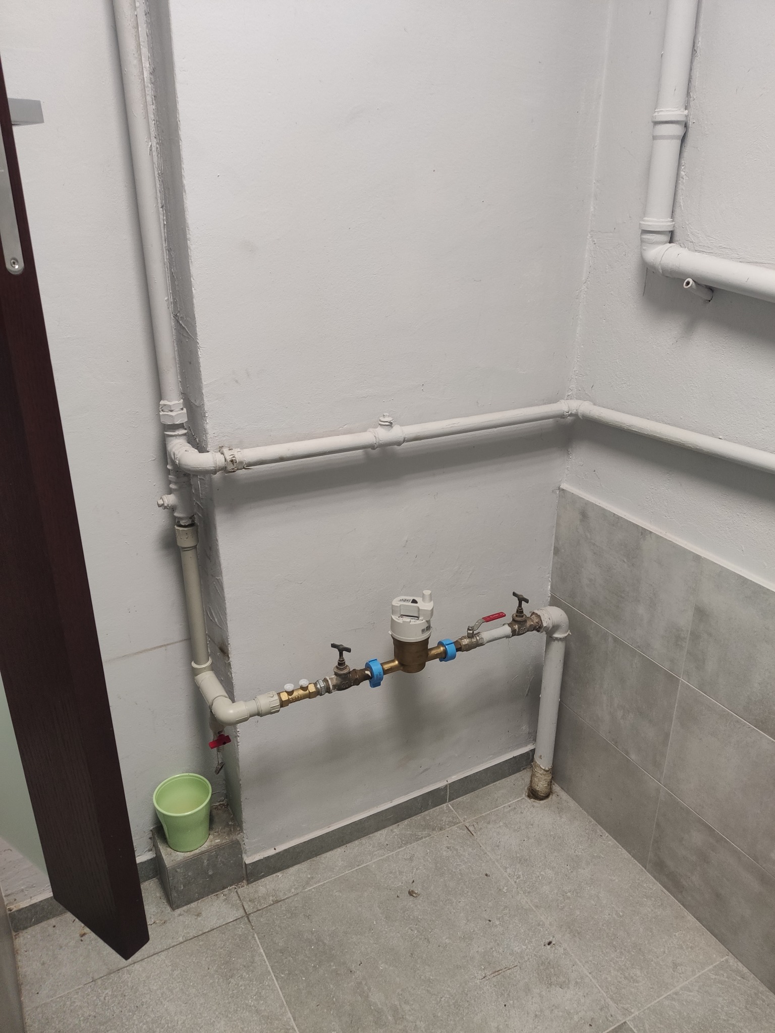 Zamontowana instalacja wodna z licznikiem w pomieszczeniu z szarą glazurą i rurami pomalowanymi na biało. Widoczny zawór i zielone wiaderko.