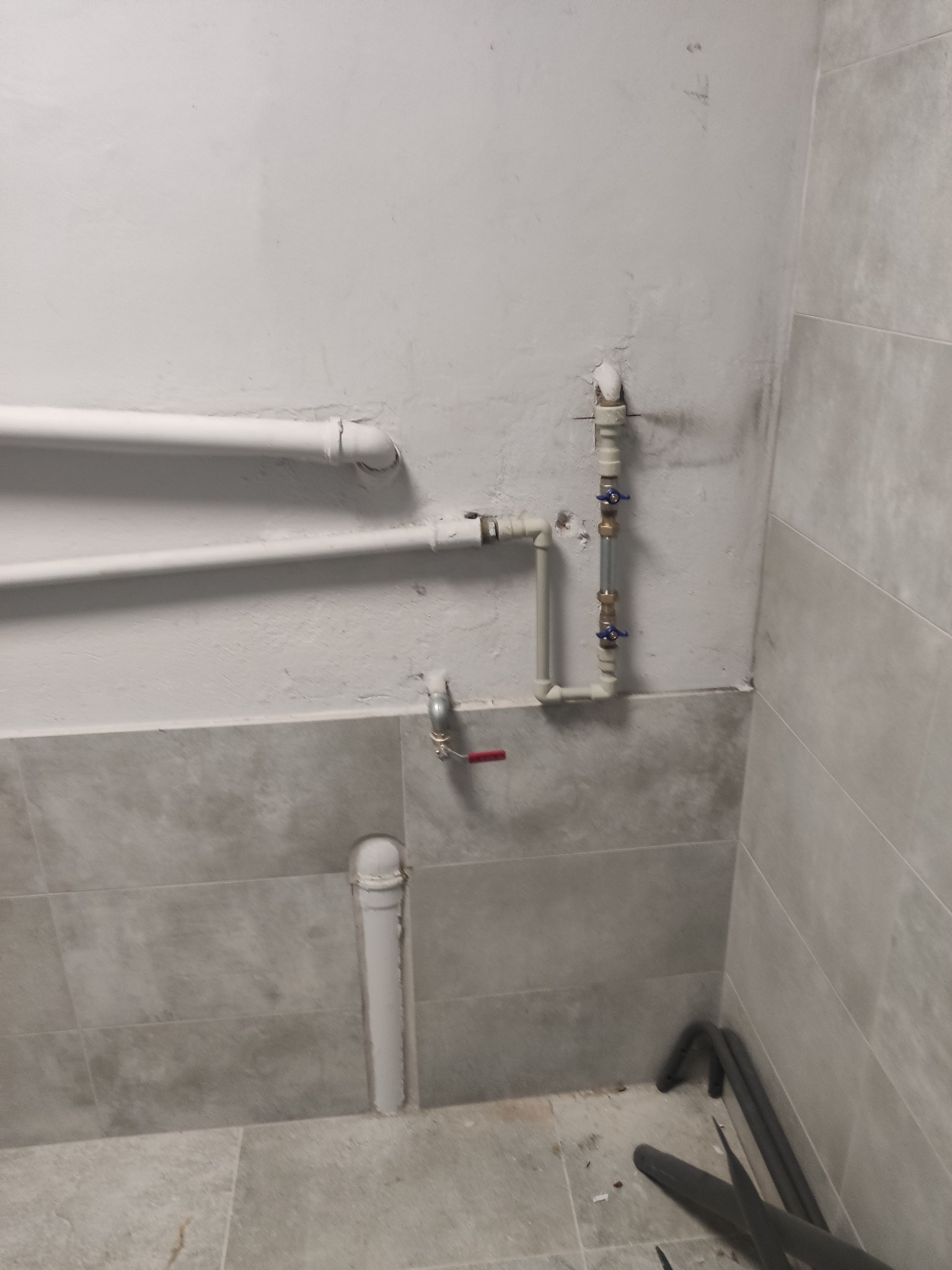 Szare płytki, białe rury i zawory w pomieszczeniu. Instalacja hydrauliczna z elementami miedzianymi i plastikowymi. Widoczne ślady po pracach instalacyjnych na ścianie.