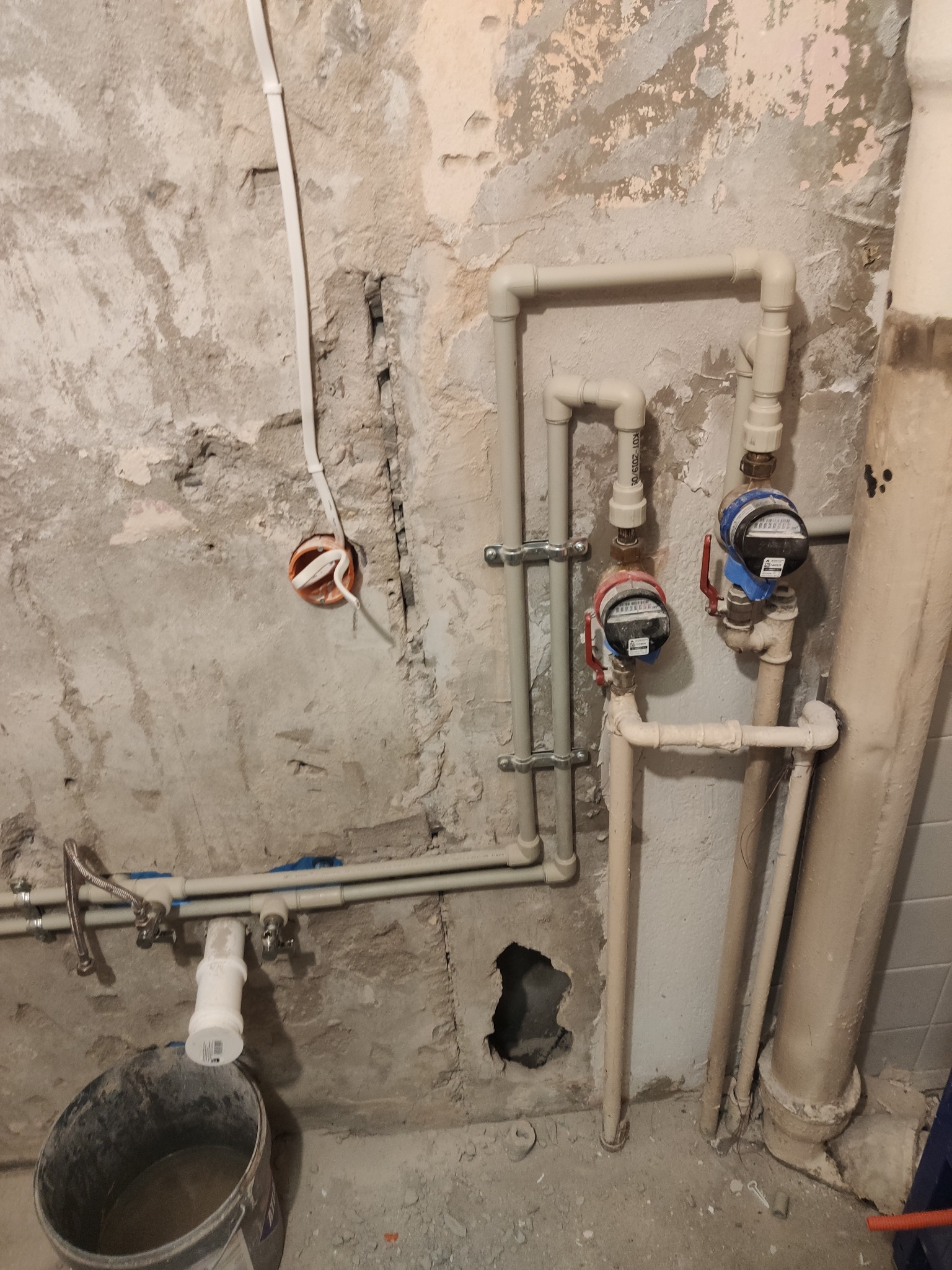 Szare rury hydrauliczne z licznikami na tle surowej ściany z widocznymi instalacjami elektrycznymi i wiadrem. Prace instalacyjne w trakcie remontu.