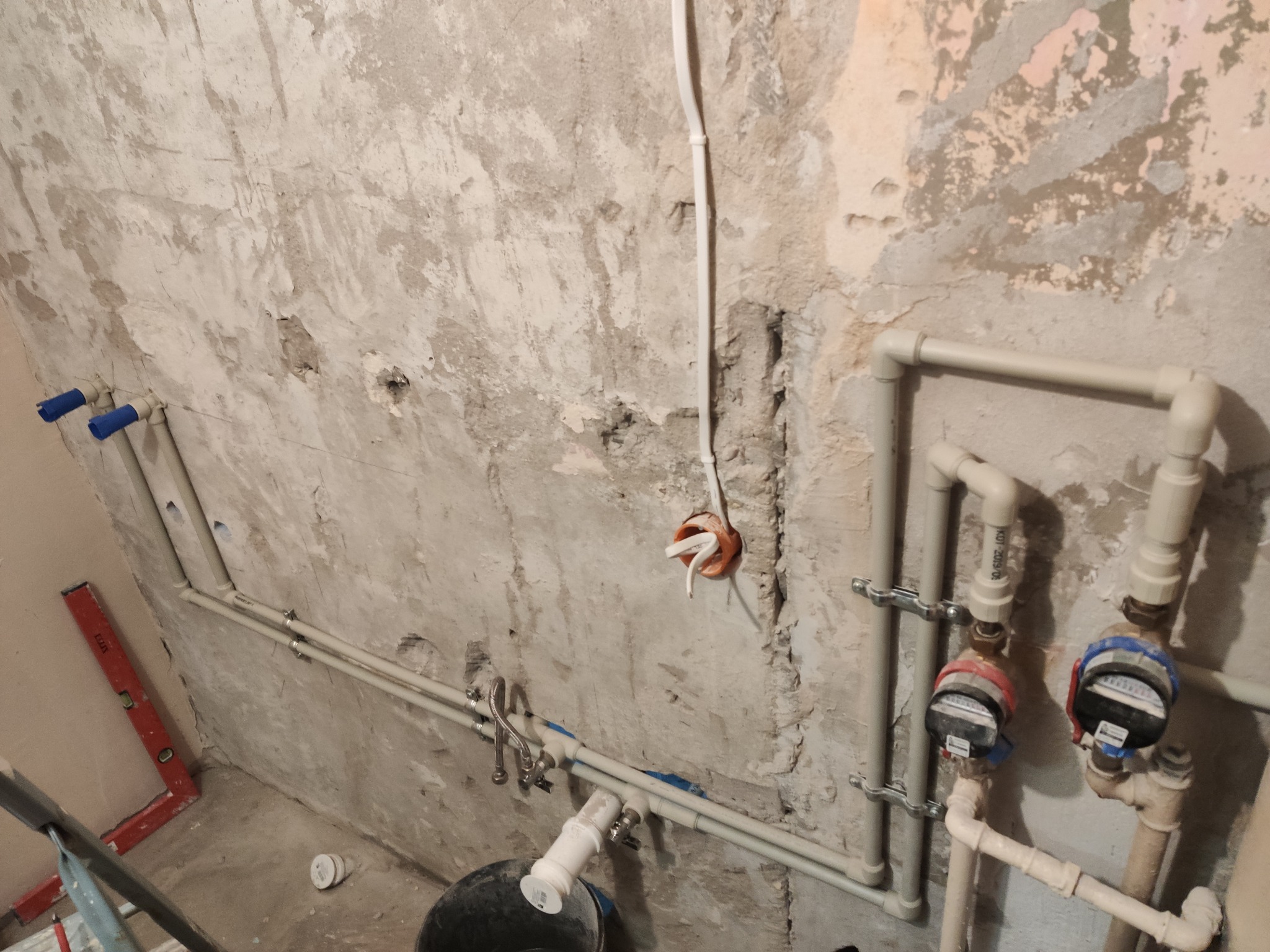 Szare ściany w trakcie remontu z widoczną instalacją hydrauliczną z rur PCV, liczniki wody i poziomica oparta o ścianę. Widoczne kable elektryczne.