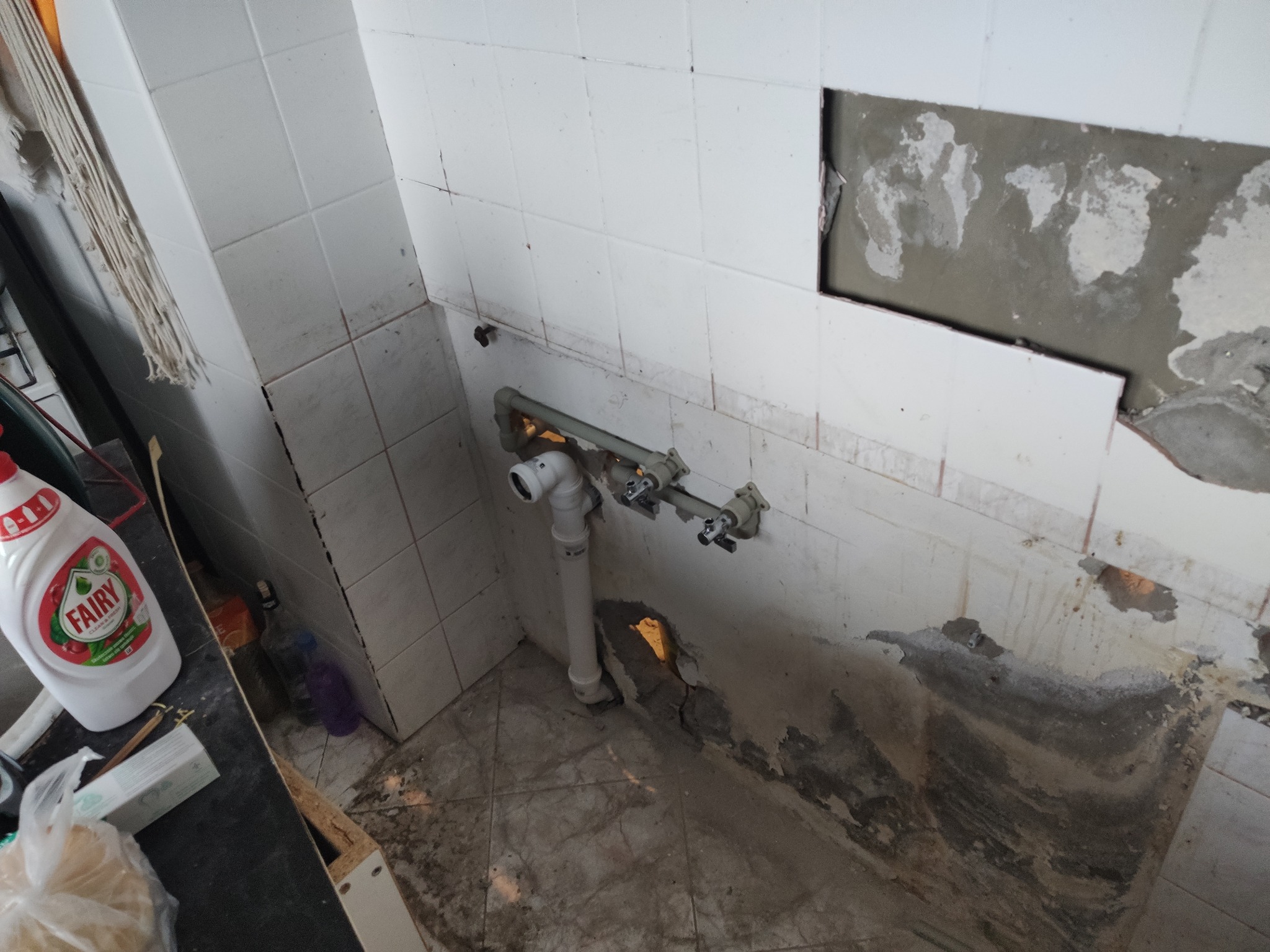 Fragment kuchni w trakcie remontu: odsłonięte rury i instalacja hydrauliczna na tle białych płytek, z widocznymi śladami po skuciu i butelką płynu do naczyń.
