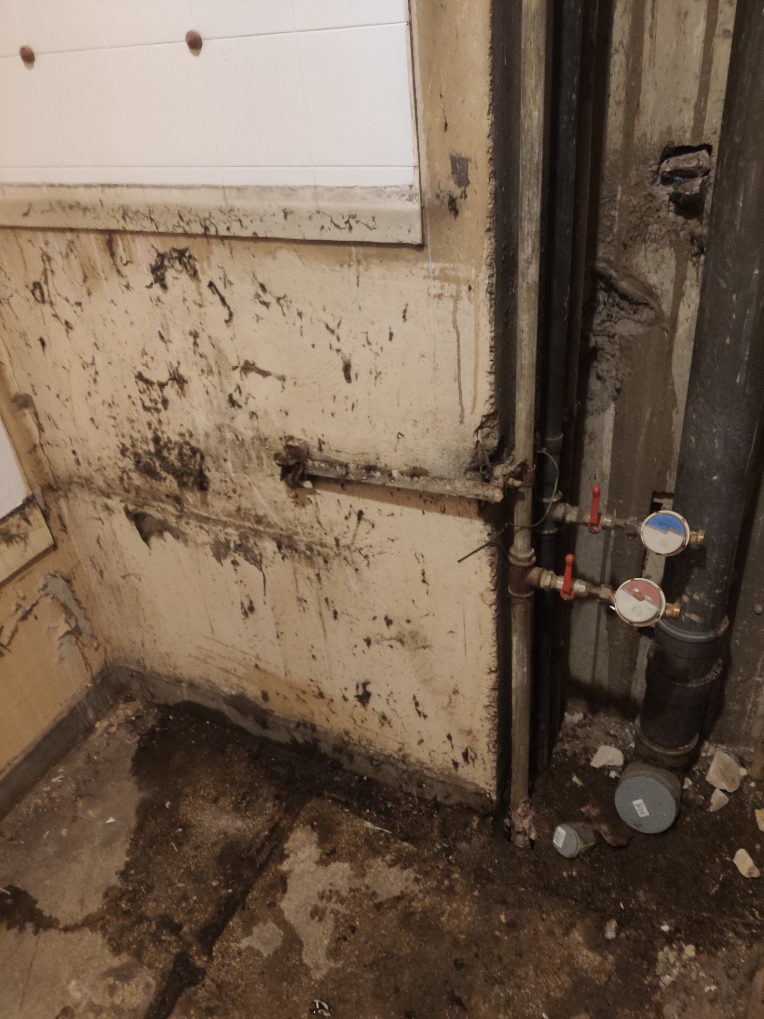 Zaniedbane pomieszczenie z widocznymi rurami, zaworami i licznikami. Ściany i podłoga zabrudzone, z widocznymi śladami wilgoci i uszkodzeń. Fragment białej glazury na ścianie.